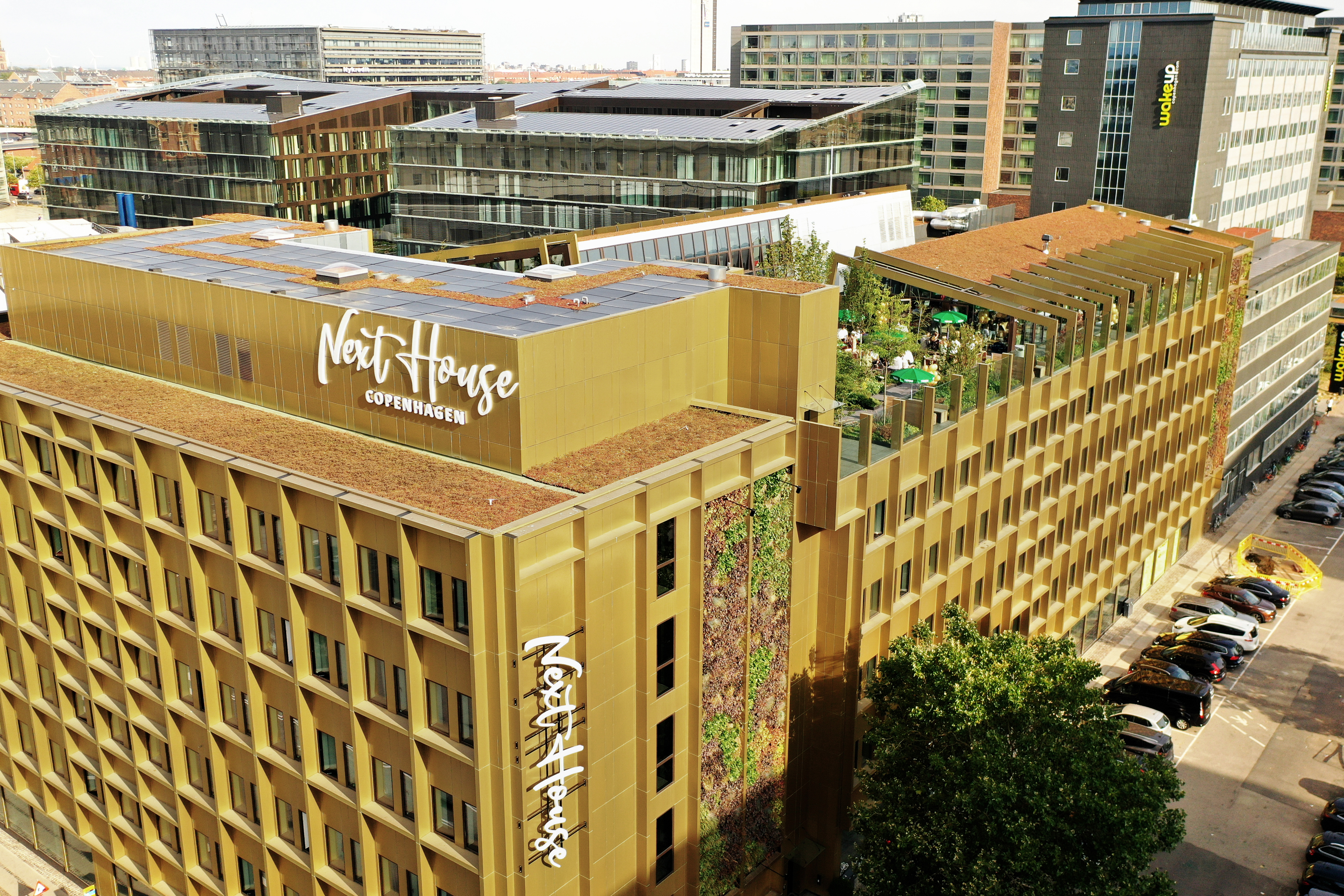 Aerial view of a modern urban area featuring buildings with green roofs. The prominent gold-colored building has Next House Copenhagen signage. In the background, multiple contemporary buildings are visible. Trees and parked cars line the streets.