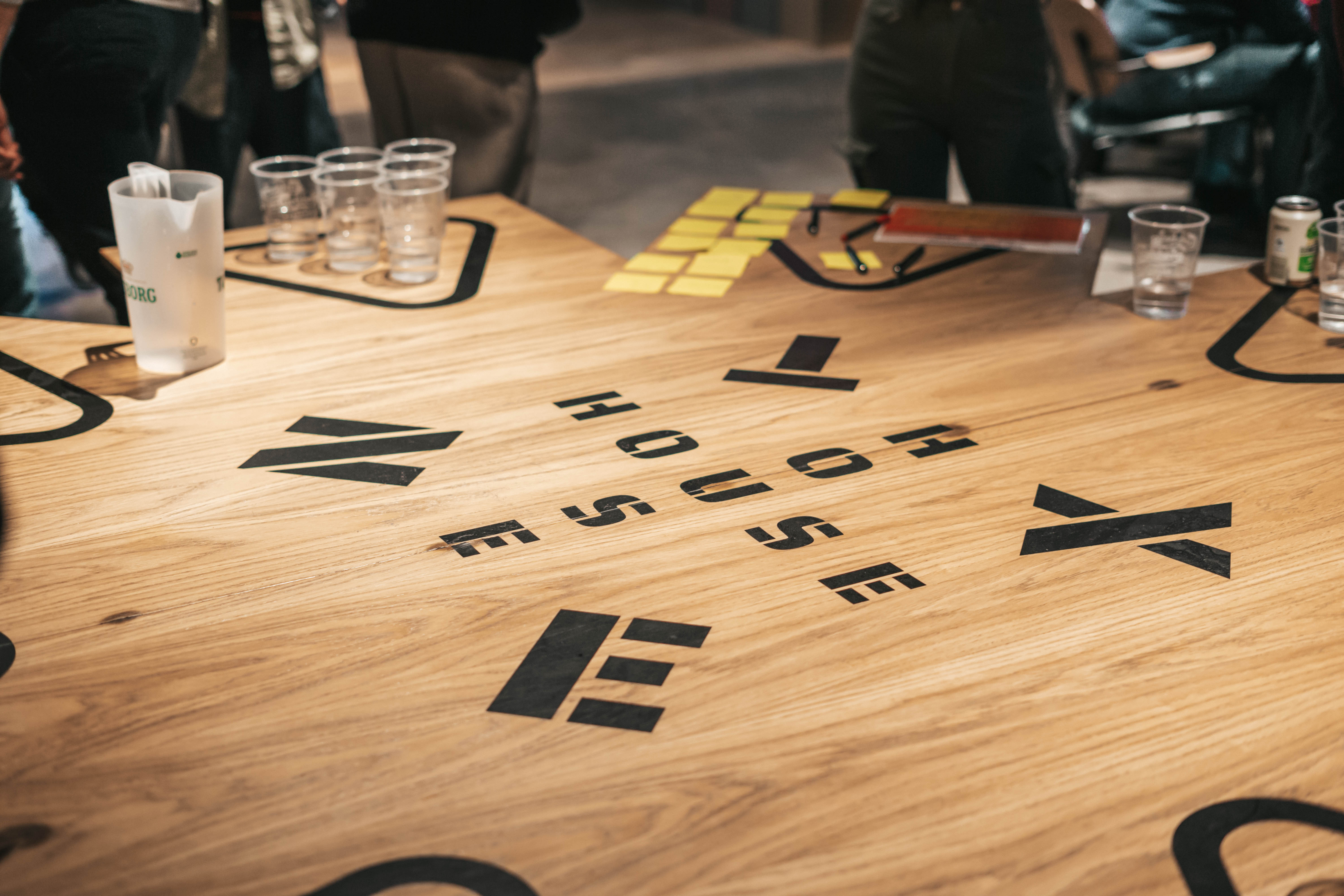 A wooden table with the words THE HOUSE written in black block letters across the surface. Empty clear cups, a pitcher, and scattered sticky notes are on the table. People are partially visible around the edges.