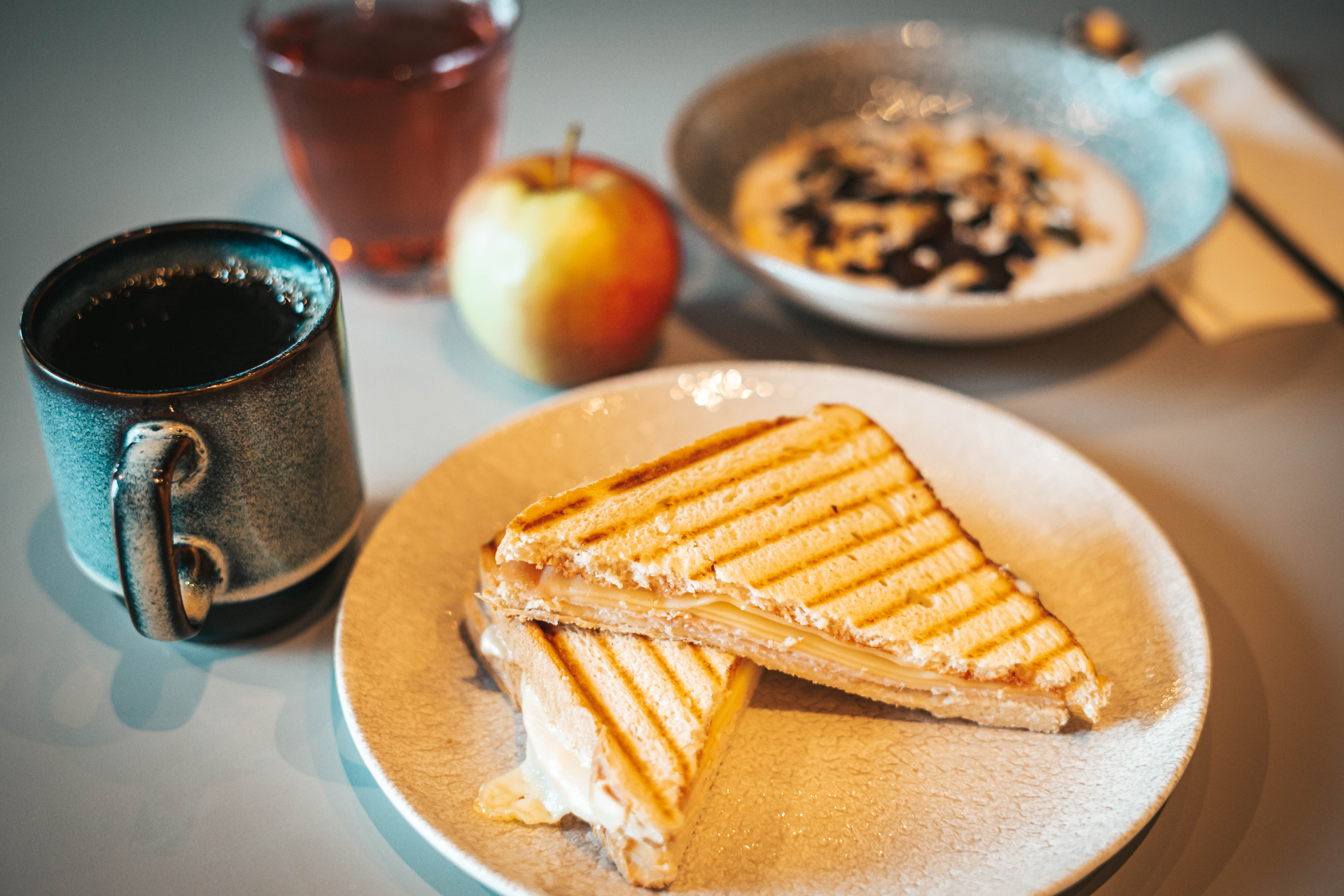 A breakfast spread featuring a plate with two grilled sandwiches, a cup of coffee, an apple, and a bowl of cereal with milk. A glass of juice is also visible in the background, all set on a light-colored table.