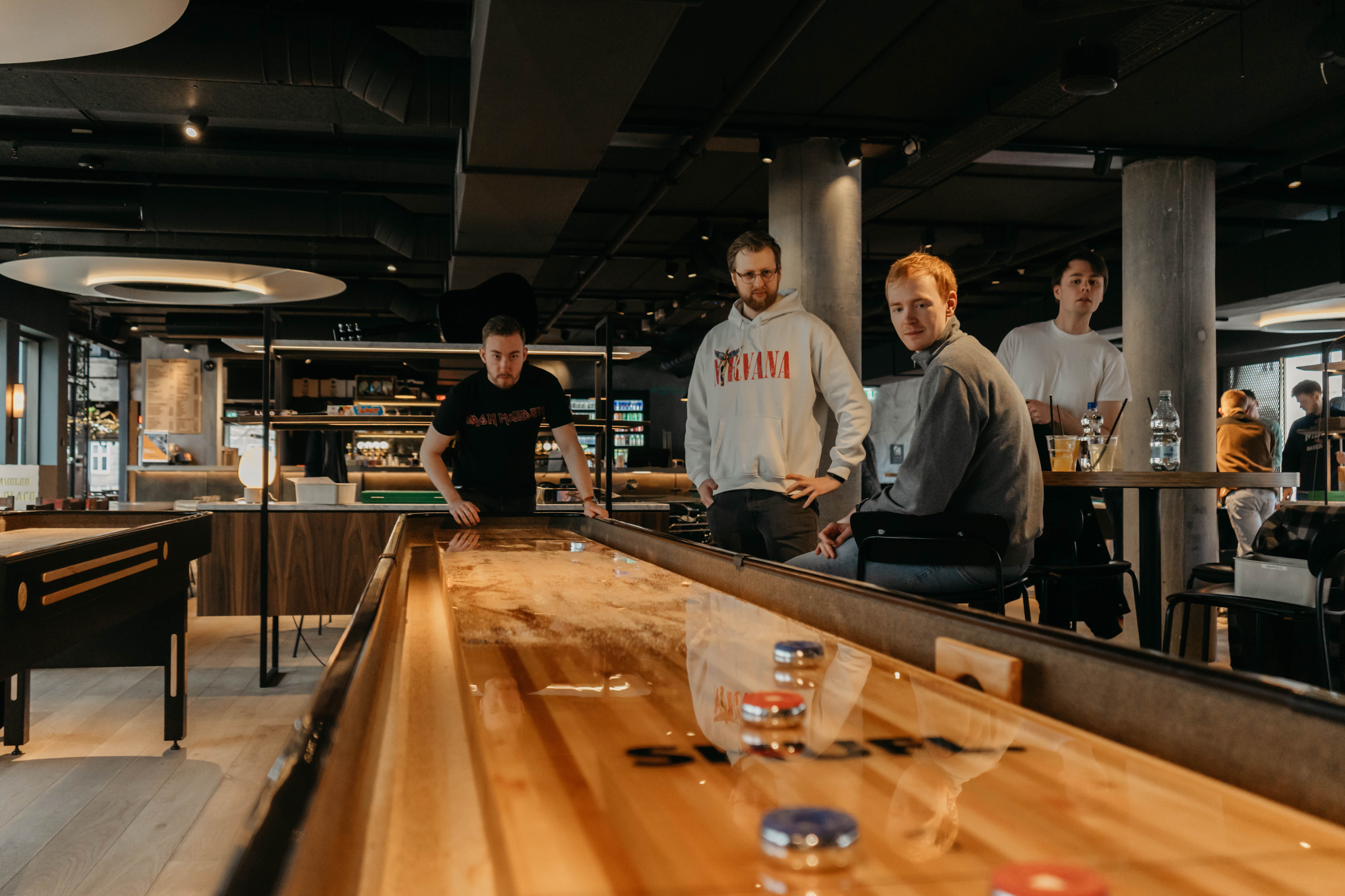 Four people are playing shuffleboard in a modern indoor space. A person on the left is taking a shot, while the others watch. The room has a stylish, warm ambiance with wooden furniture and low lighting.