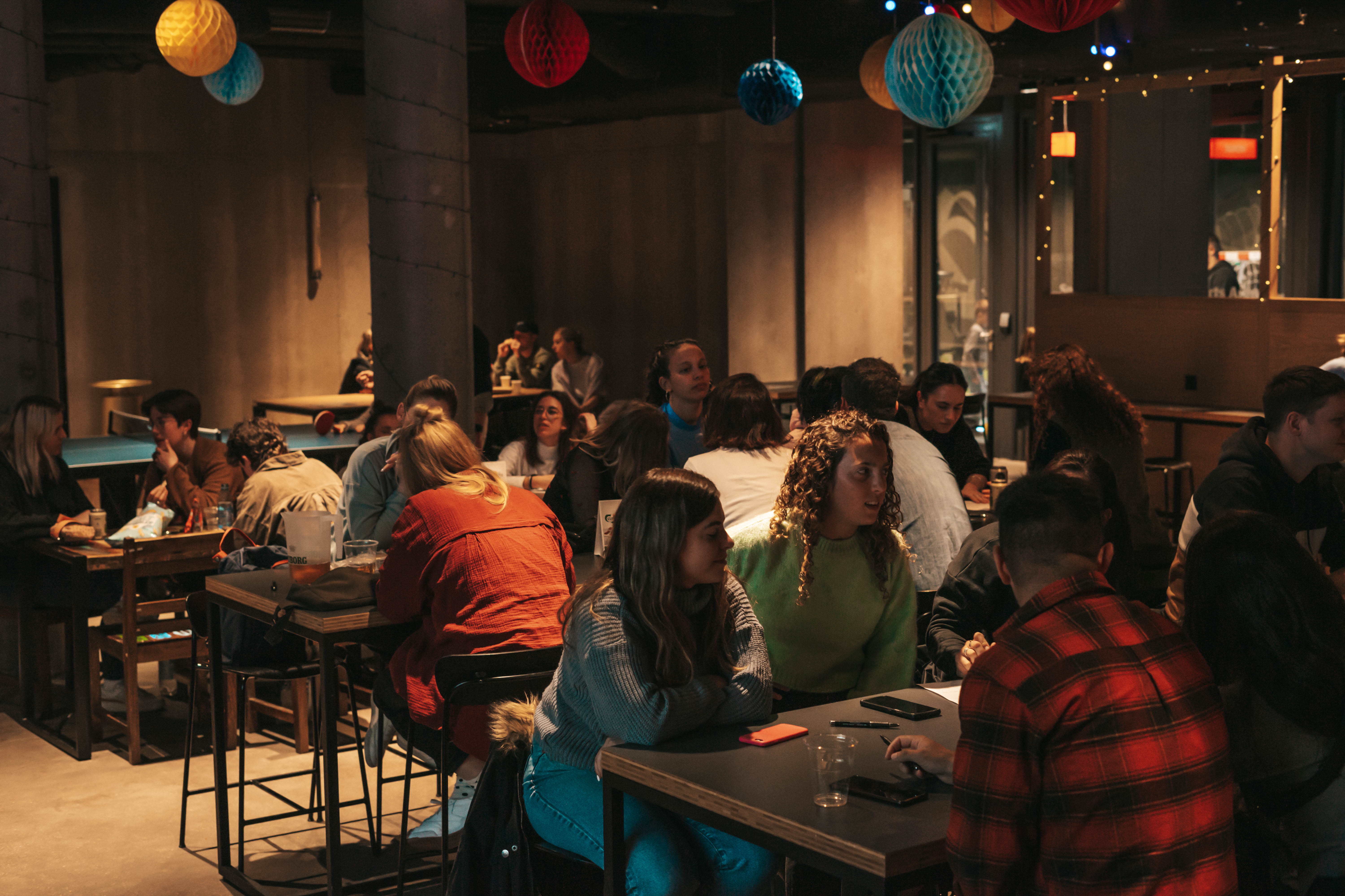 A dimly lit indoor café filled with people sitting and chatting at tables. Festive paper lanterns hang from the ceiling, creating a cozy atmosphere. Warm, ambient light casts a soft glow over the group, enhancing the intimate setting.