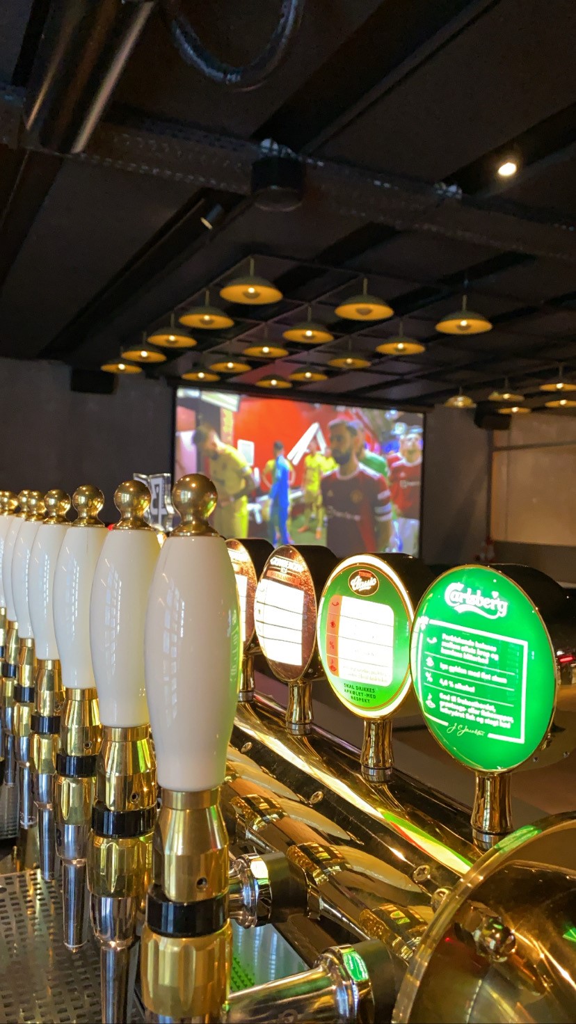 A row of draft beer taps in a dimly lit bar, with a large screen in the background showing a sports game broadcast. The ceiling has round, hanging lights, and the taps are shiny with white handles.