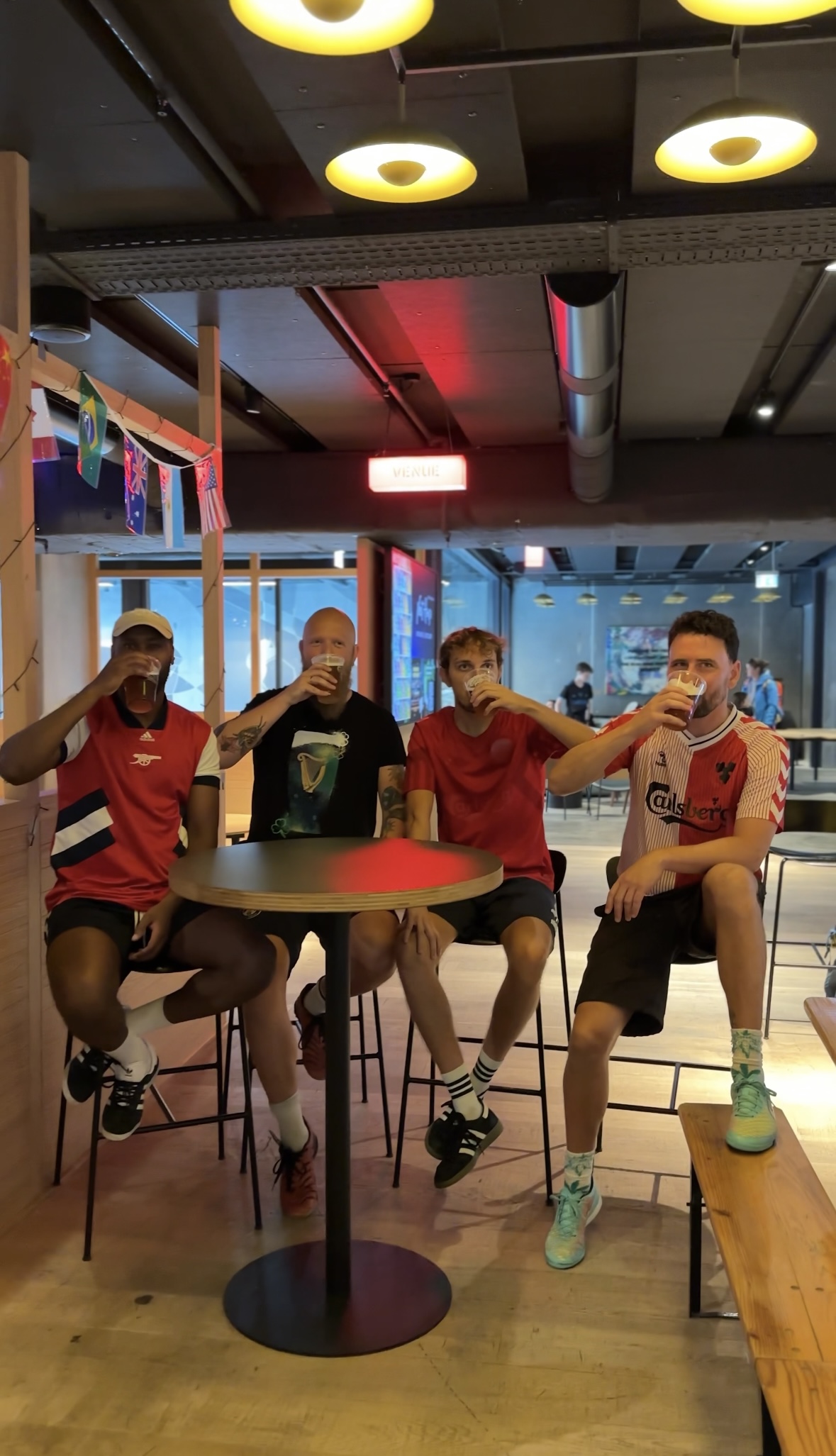 Four people sit at a high table in a bar, each holding a drink. The group includes two wearing red shirts and one with a black top. Flags hang as decorations in the background, and the setting is casual with wooden seats and overhead lights.