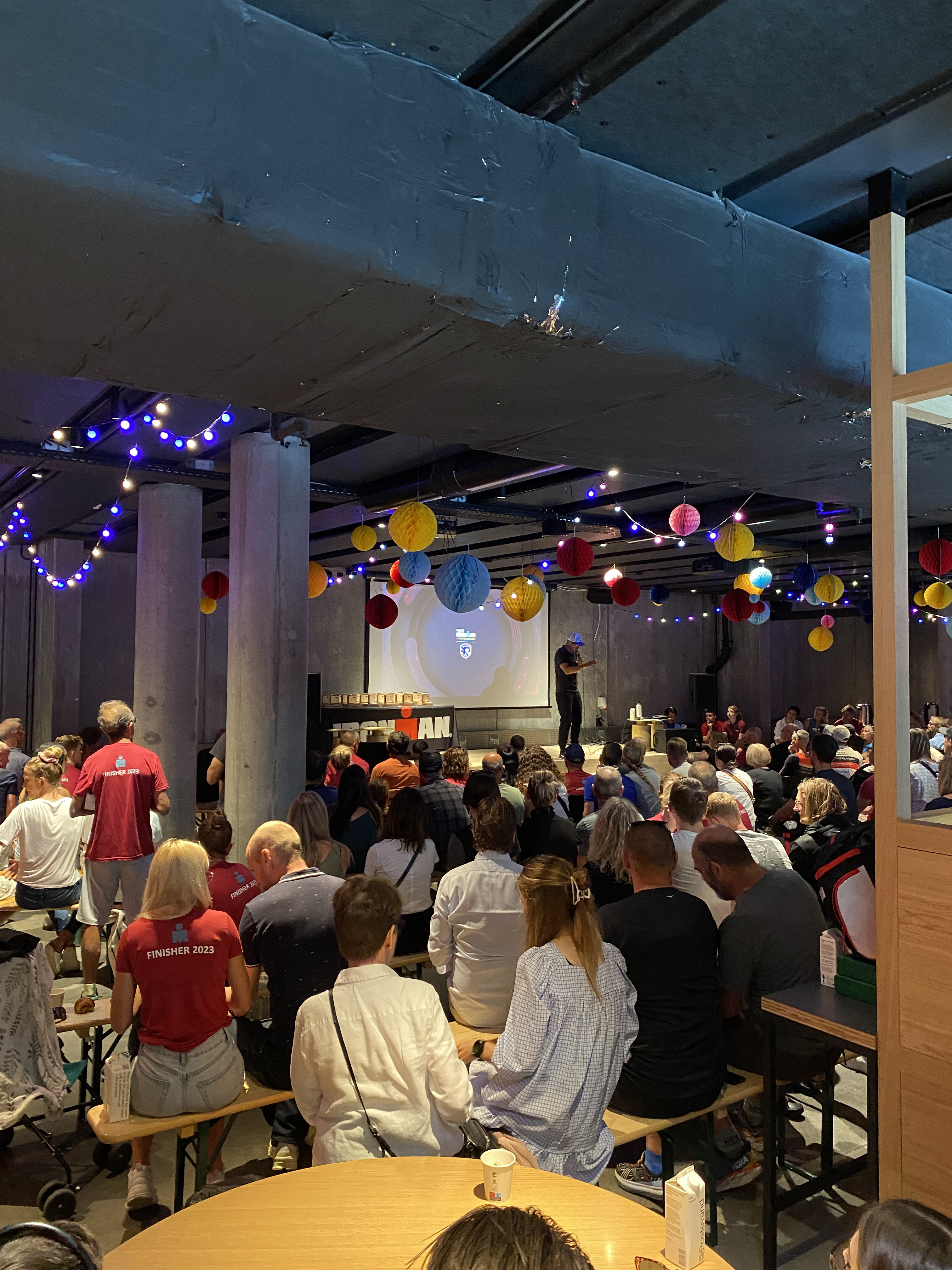 A large group of people seated indoors, attending a presentation. The room is decorated with colorful hanging lights and lanterns. A person stands at the front near a projecting screen displaying a slide.