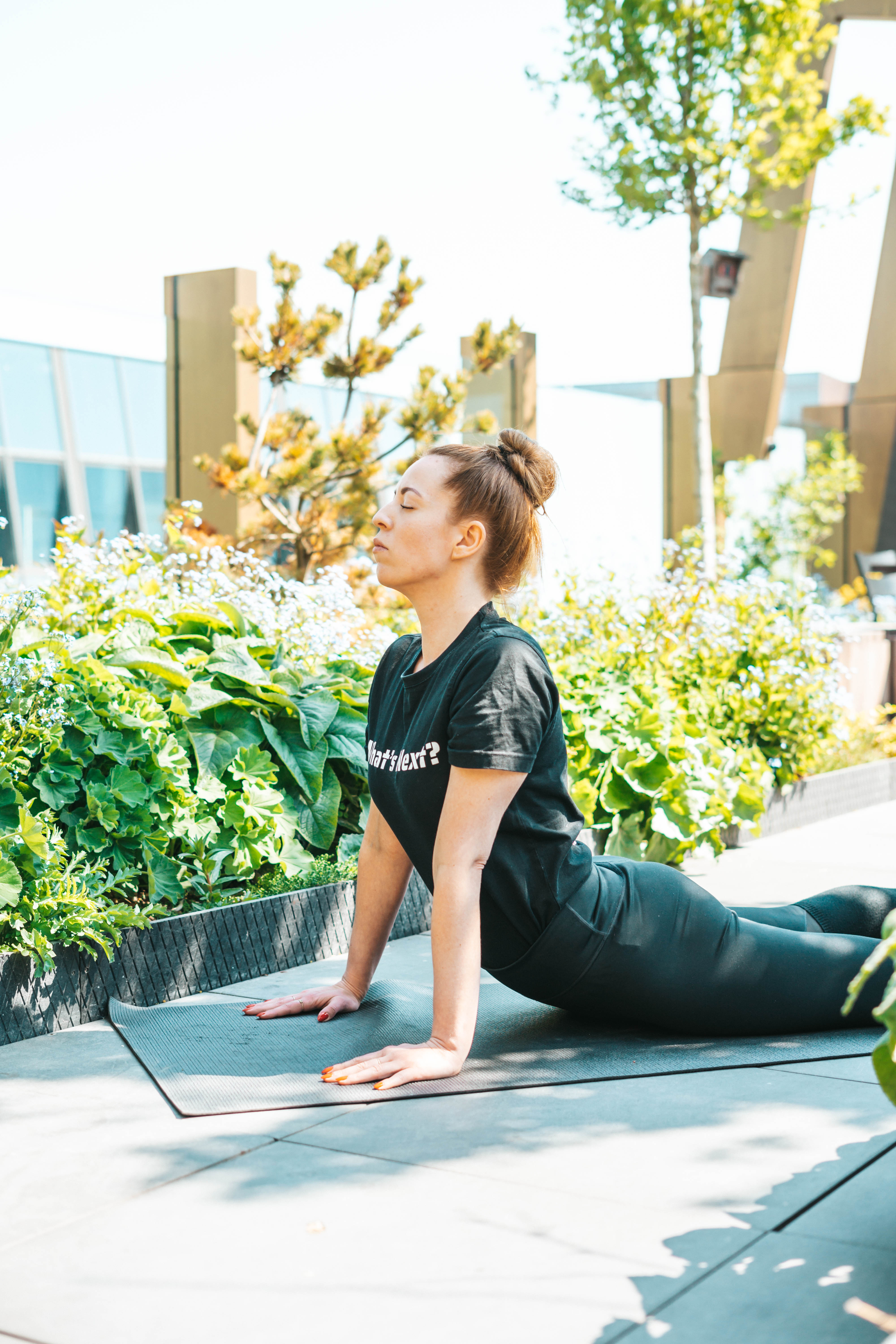 A person in a black outfit performs a yoga pose on a mat outdoors. Theyre in a garden with lush greenery and sunlight filtering through. The surroundings suggest a peaceful, natural environment.