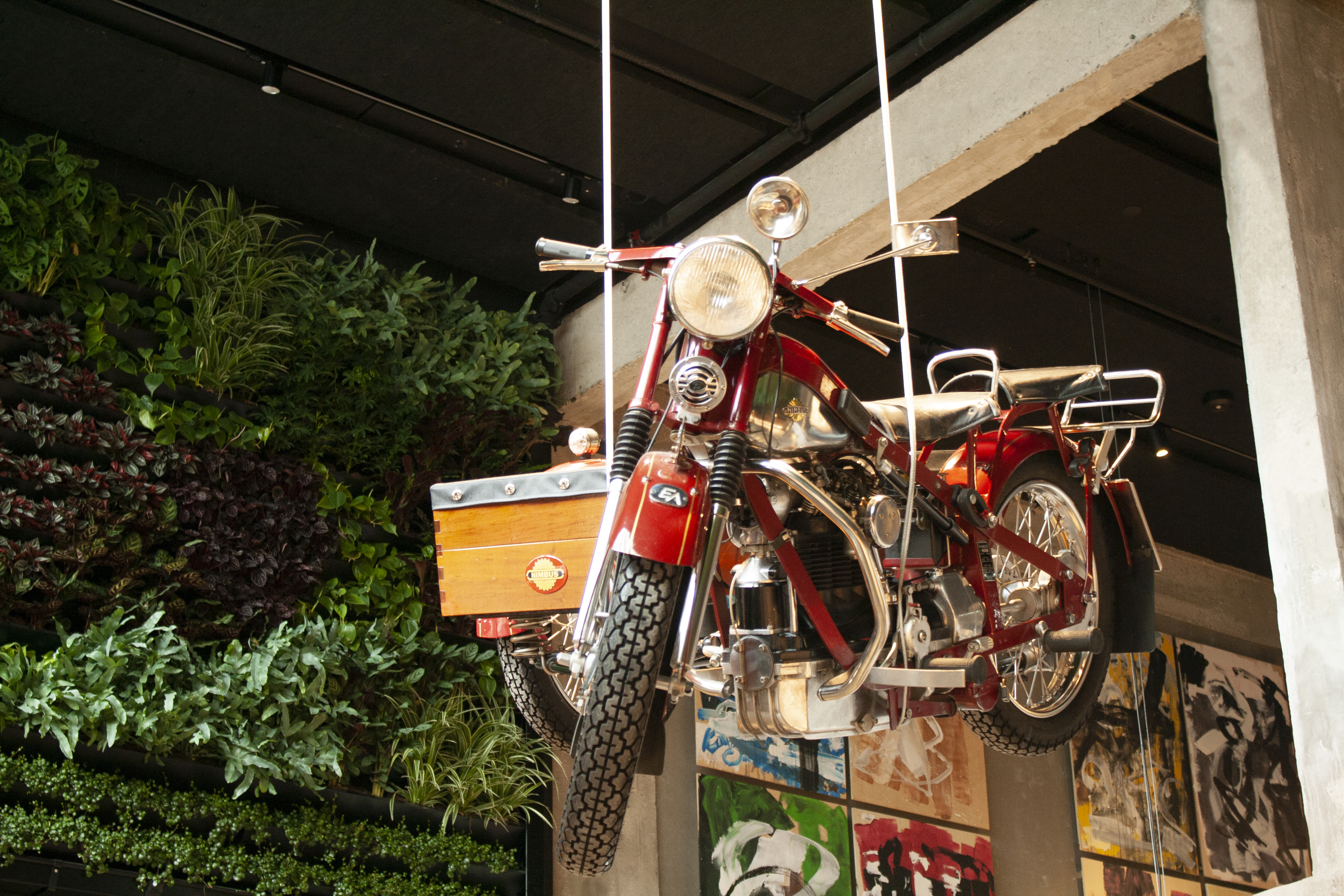 A vintage red motorcycle suspended from the ceiling, with greenery and colorful artwork on the wall behind it. The handlebars are shiny, and the motorcycle features a wooden cargo compartment.
