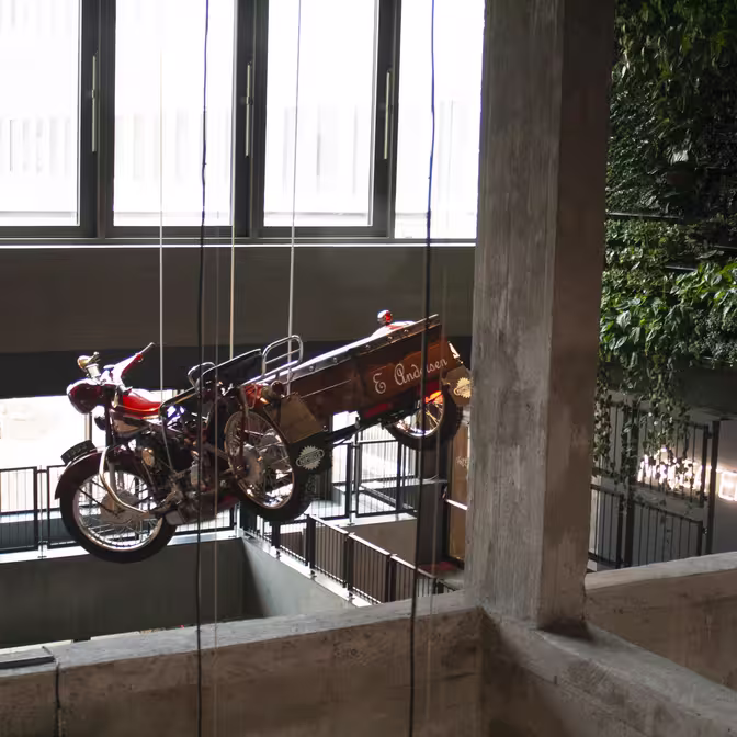 En motorcykel med sidevogn er ophængt af kabler i et interiør i industriel stil med store vinduer og en plantedækket væg. Lyset filtrerer gennem vinduerne og skaber en lys og luftig atmosfære.