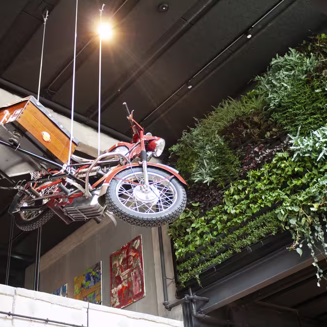 En vintage motorcykel med sidevogn hænger ophængt i et moderne interiør. Til højre dækker en lodret have med frodige grønne planter en mur. Kraftig overhead-belysning oplyser scenen med kunstværker synlige på en nærliggende væg.