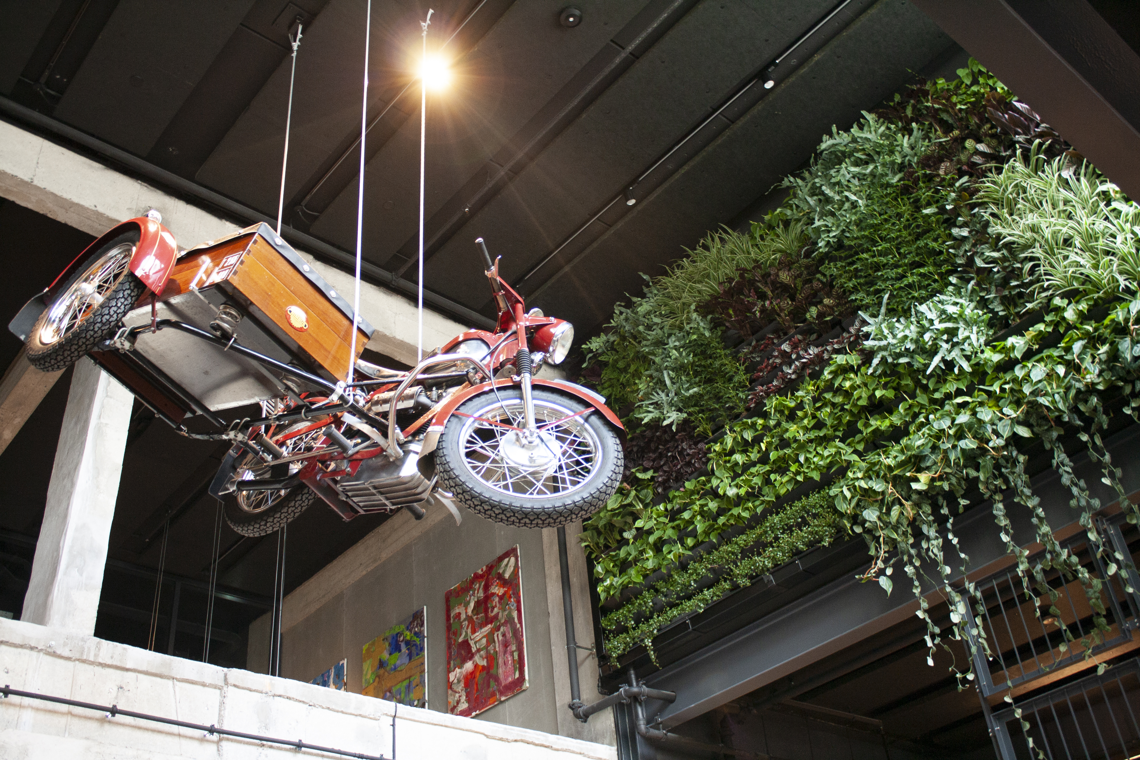 An interior shot of a room featuring a red vintage motorcycle with a sidecar suspended from the ceiling. The wall behind it is adorned with vibrant vertical greenery. Artwork is displayed on the adjacent wall, and a spotlight illuminates the motorcycle.