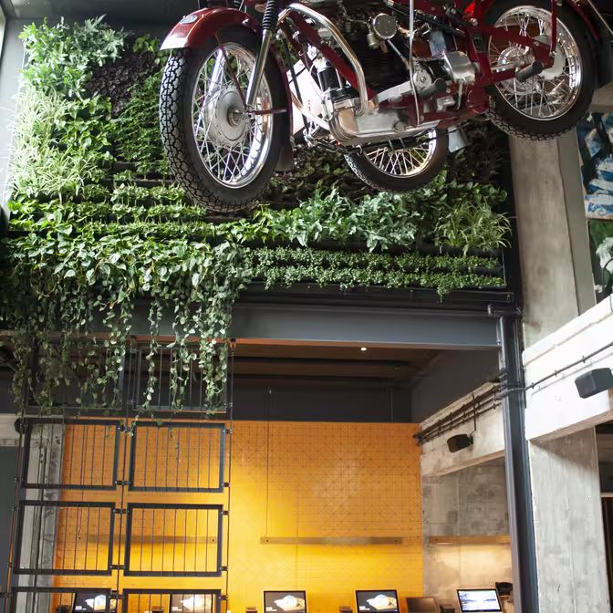 En vintage rød motorcykel er ophængt fra loftet i et moderne interiør. Nedenfor tilføjer en lodret havevæg grønt, og flere bærbare computere vises på en lang træbord mod en tekstureret gul væg.