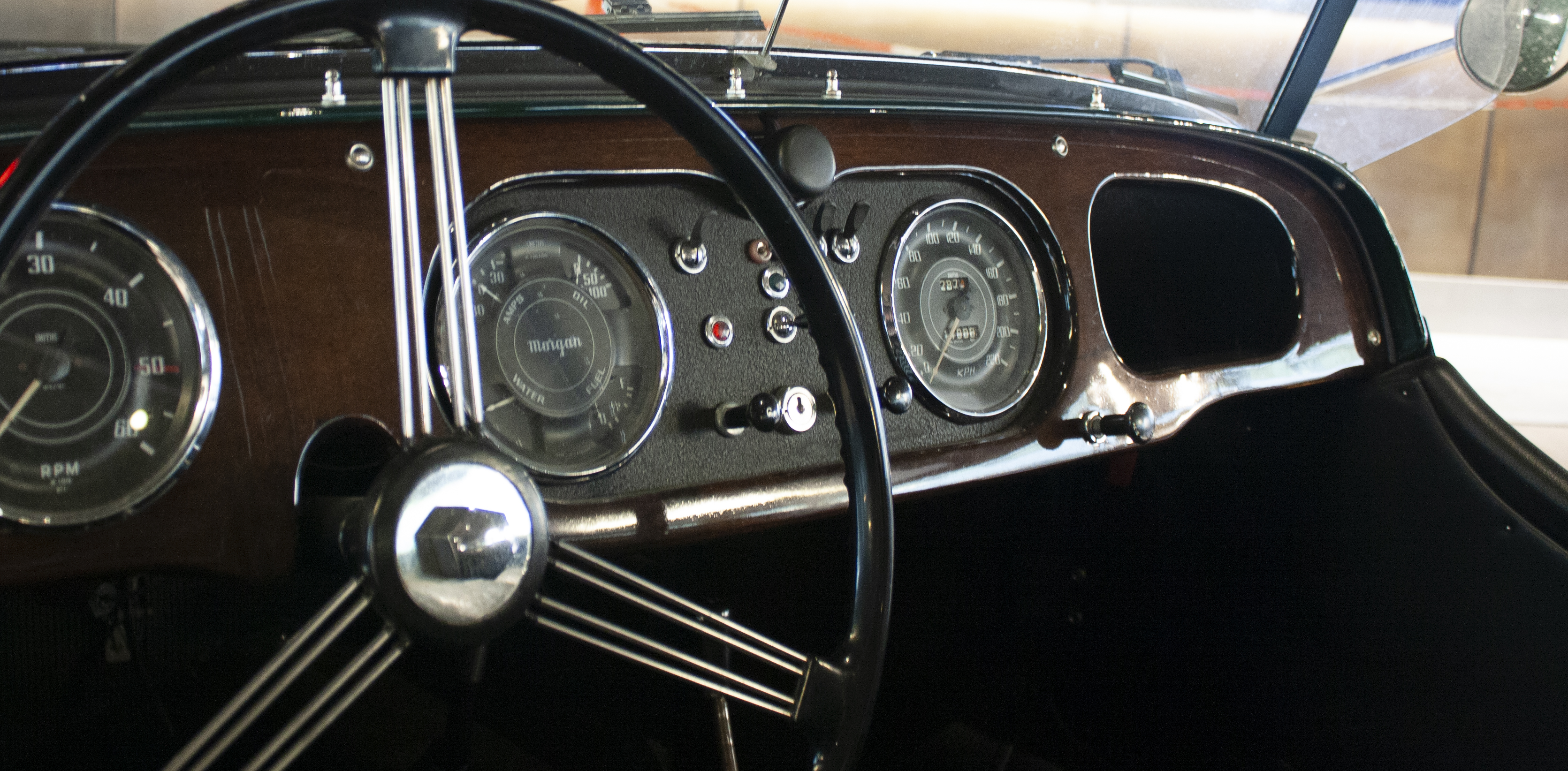 Close-up of a vintage car dashboard featuring a classic steering wheel, speedometer, and various gauges set against a wooden panel. The interior has a retro design with chrome accents and a historical automotive feel.