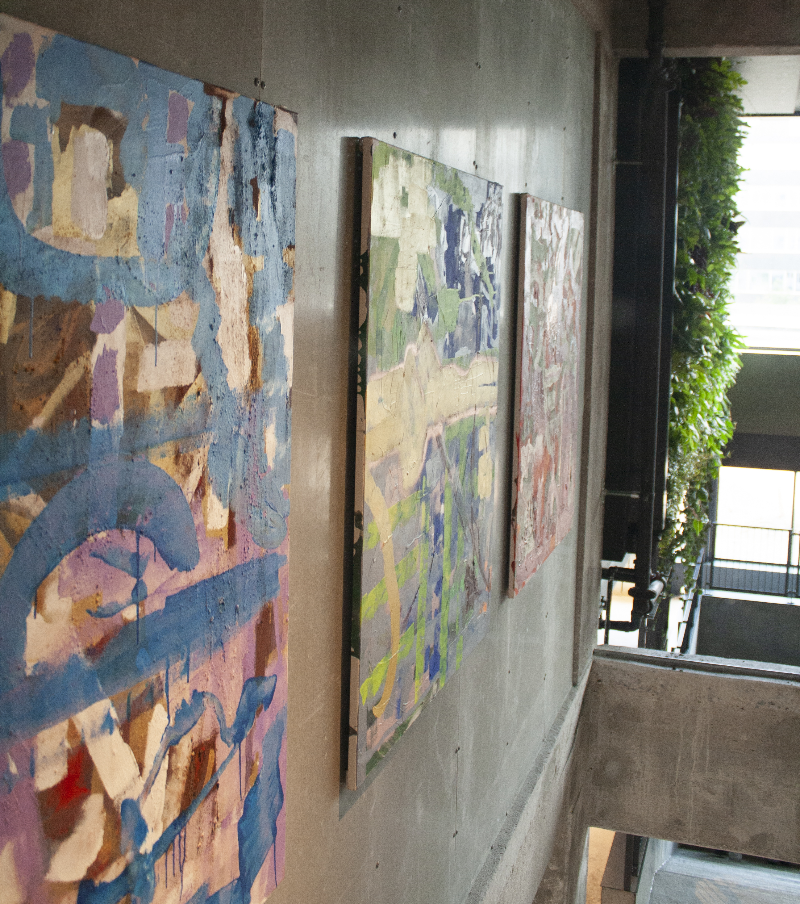 Abstract paintings with various colors hang on a concrete wall in a modern building with abundant greenery and glass railings. Sunlight filters through large windows, highlighting the mix of art and architecture.