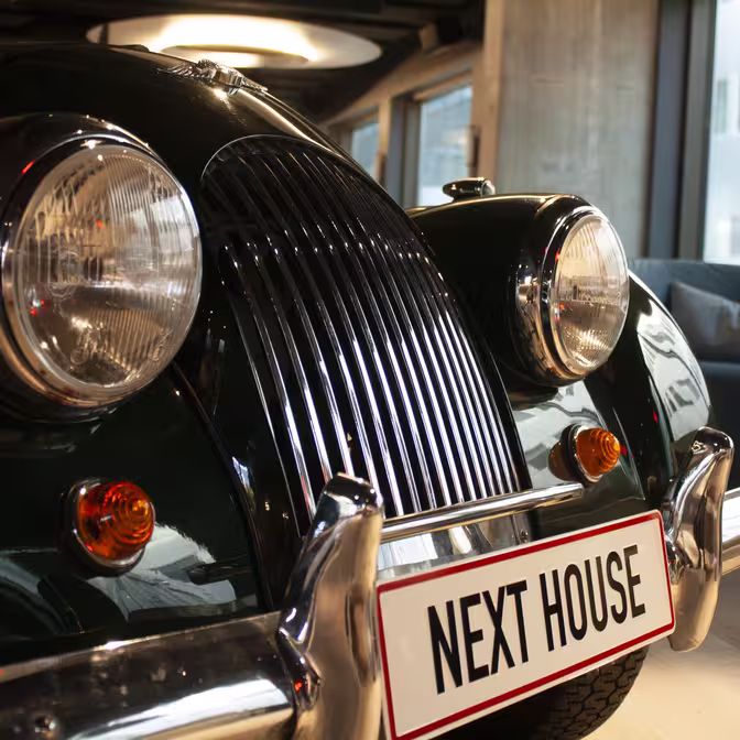 Et nærbillede af en klassisk mørkegrøn bil med et skinnende kromgitter og runde forlygter. På den forreste nummerplade står der "NEXT HOUSE." Bilen er indendørs, med moderne bygningsvinduer synlige i baggrunden.