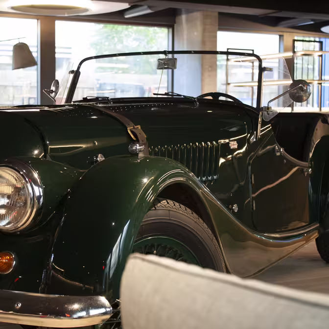 En klassisk mørkegrøn cabriolet er parkeret indendørs under blød belysning. Veteranbilen har en krom kofanger, runde forlygter og en slank, skinnende krop. Der er moderne arkitektoniske elementer og møbler synlige i baggrunden.