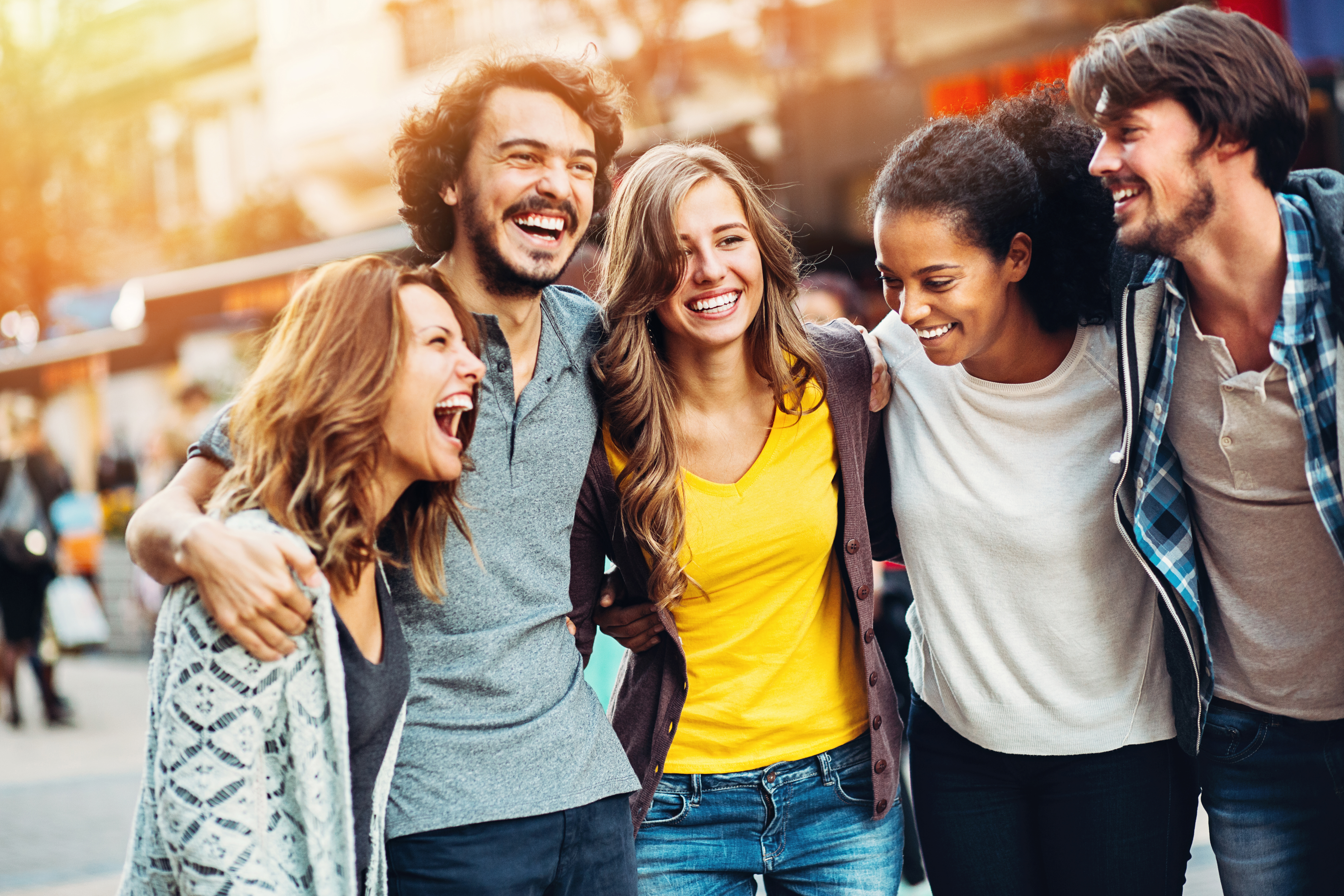 A group of five friends stand closely and smile joyfully outdoors. They are casually dressed, with arms around each other. The background shows a blurred street scene with warm lighting, suggesting a lively and cheerful urban environment.