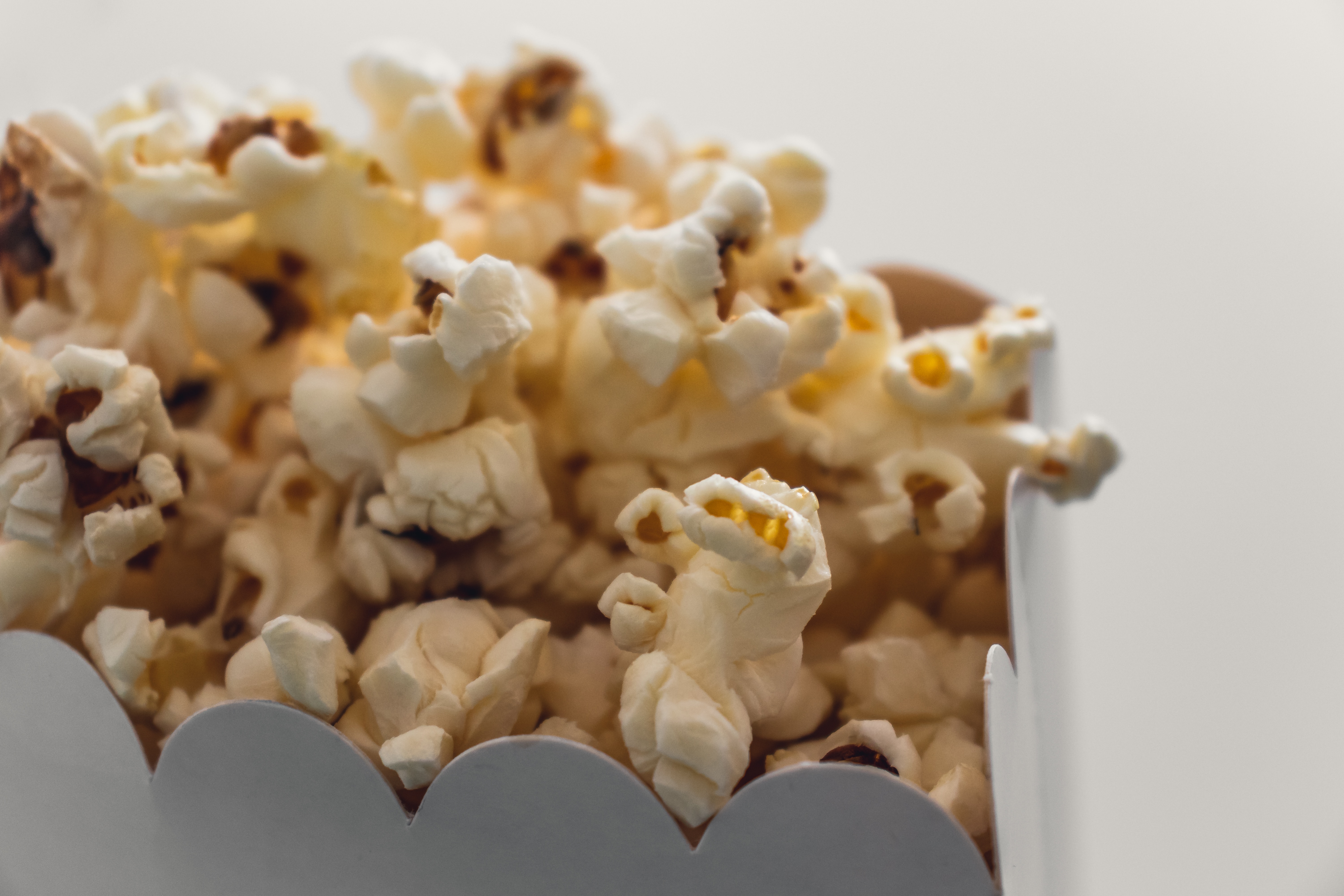 Close-up of freshly popped popcorn in a white, scalloped-edge box against a white background. The popcorn is fluffy and golden, with some kernels visible.