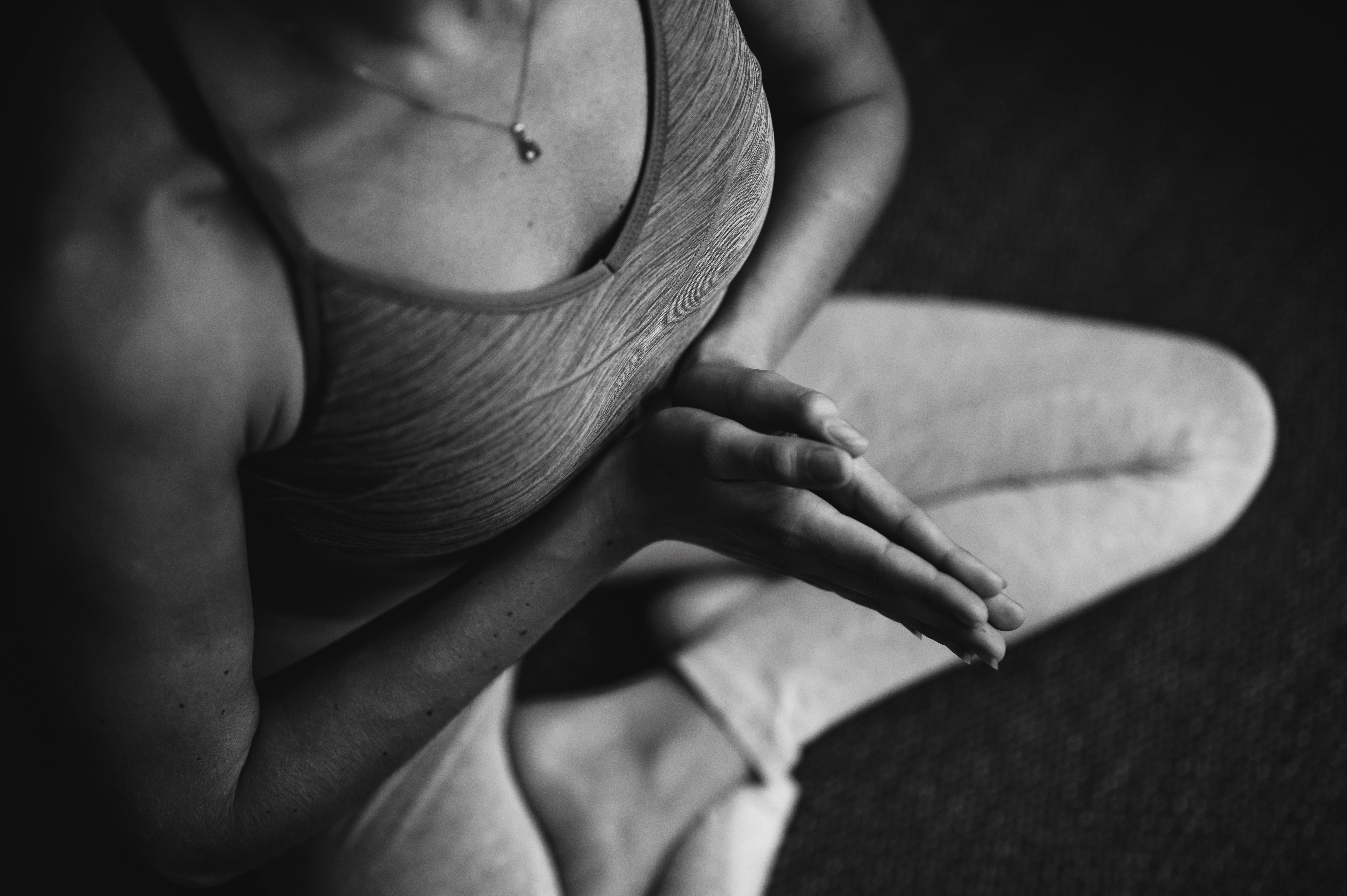 Black and white image of a person sitting cross-legged, wearing a tank top and leggings. Their hands are pressed together in a meditative pose. Only the upper torso and partial legs are visible.