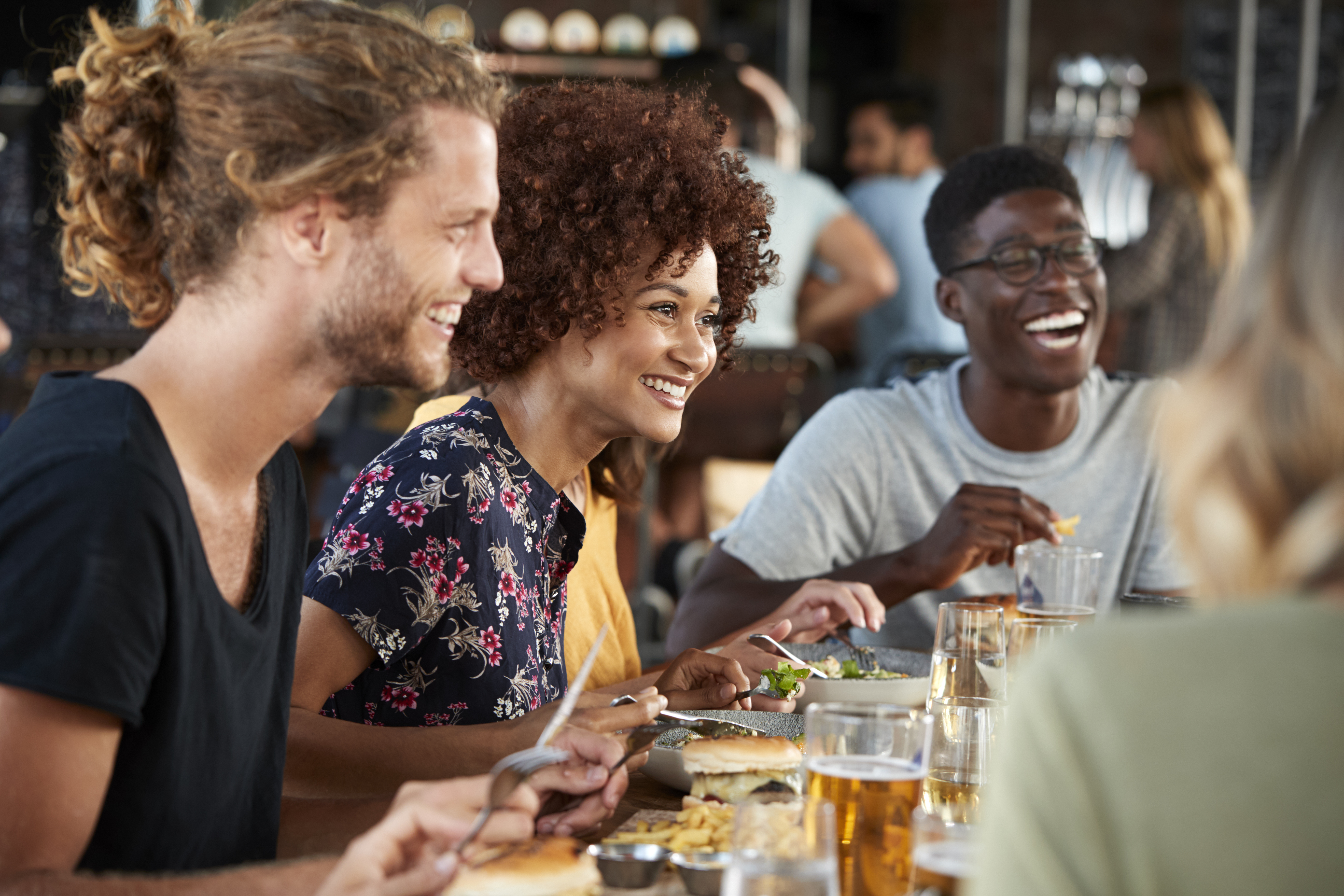 A group of four friends sitting at a restaurant table, laughing and enjoying a meal together. They have plates of food and glasses of drinks in front of them. The atmosphere appears lively and cheerful.