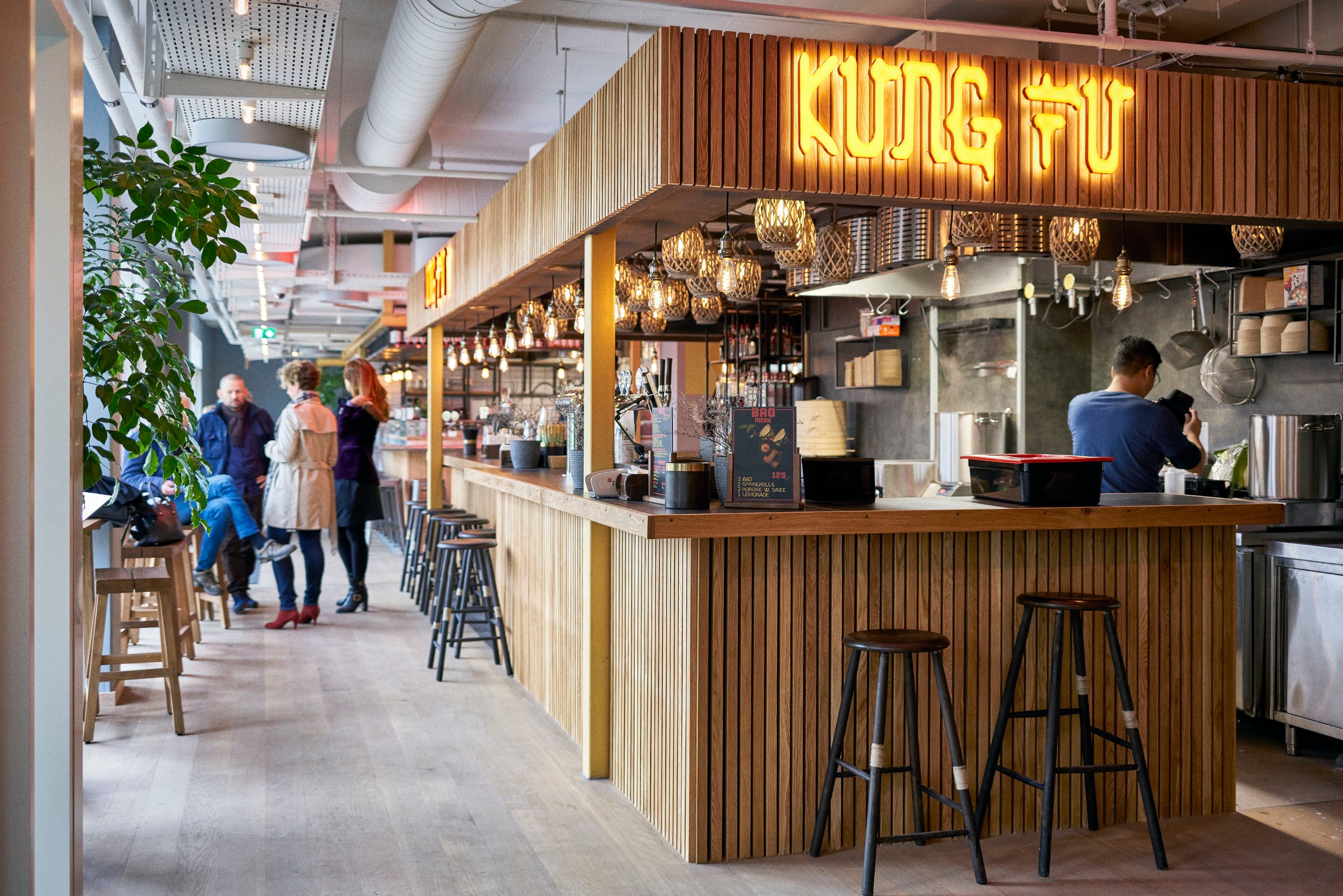 A modern restaurant interior with wooden accents and a glowing KUNG FU sign above the counter. Several people are casually interacting near the counter, while a person prepares food in the kitchen. Hanging lights add a warm ambiance.