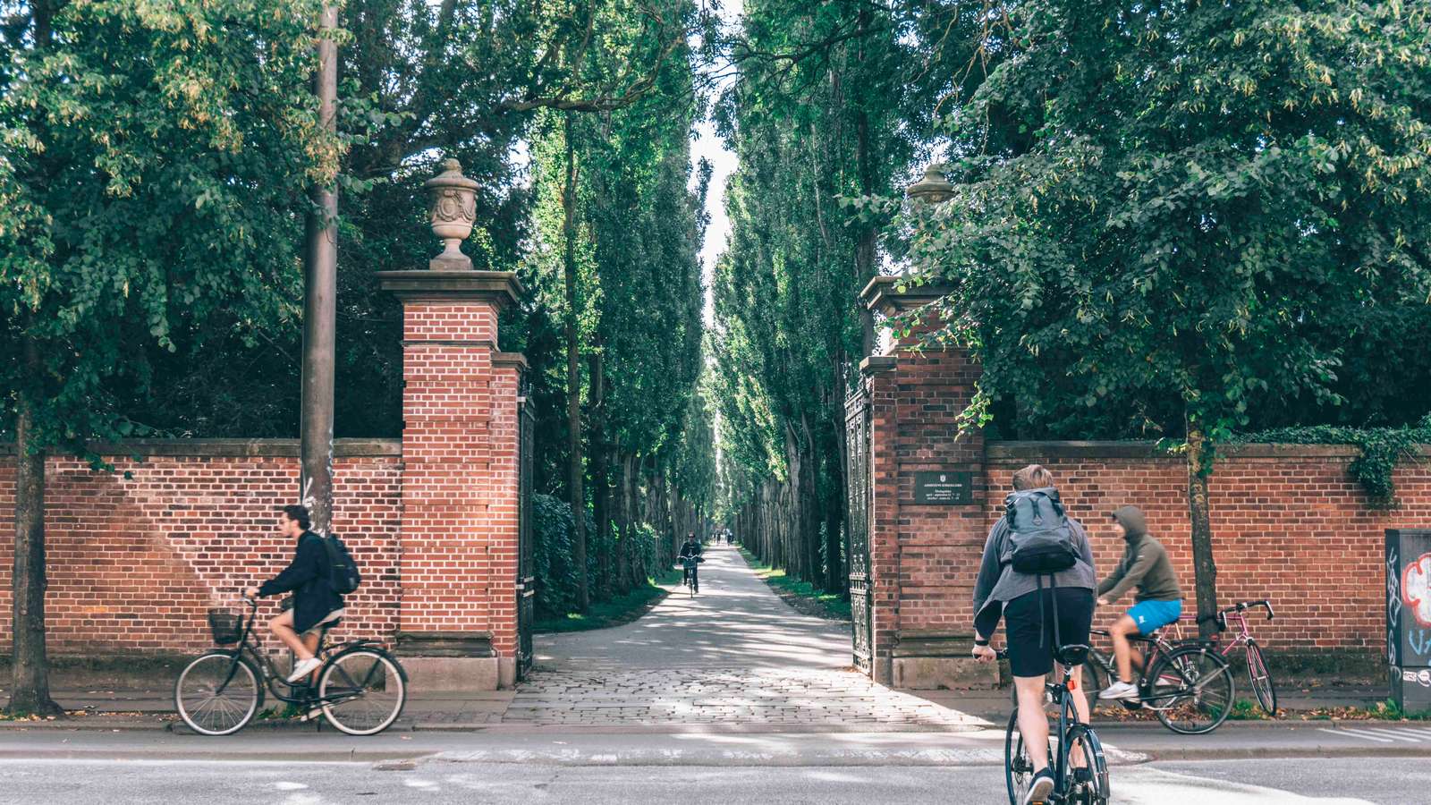 Two cyclists ride past a red brick entrance flanked by tall hedges and trees, leading to a shaded pathway. The scene captures a serene and green urban space under a clear sky.