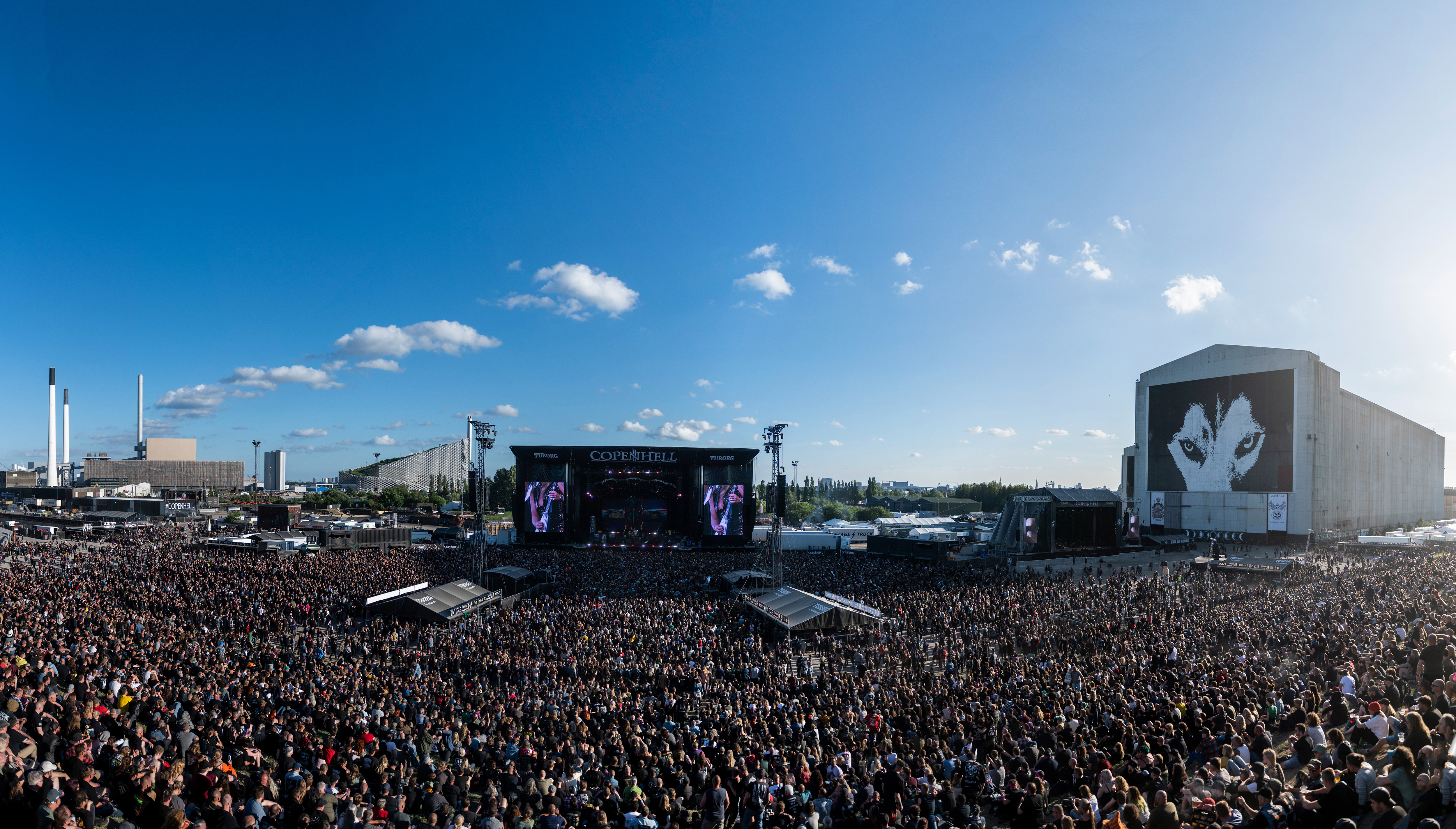 A large crowd at an outdoor music festival under a clear blue sky. The stage displays a band performing, and theres a large mural of a face on a nearby building. Industrial structures are visible in the distance.