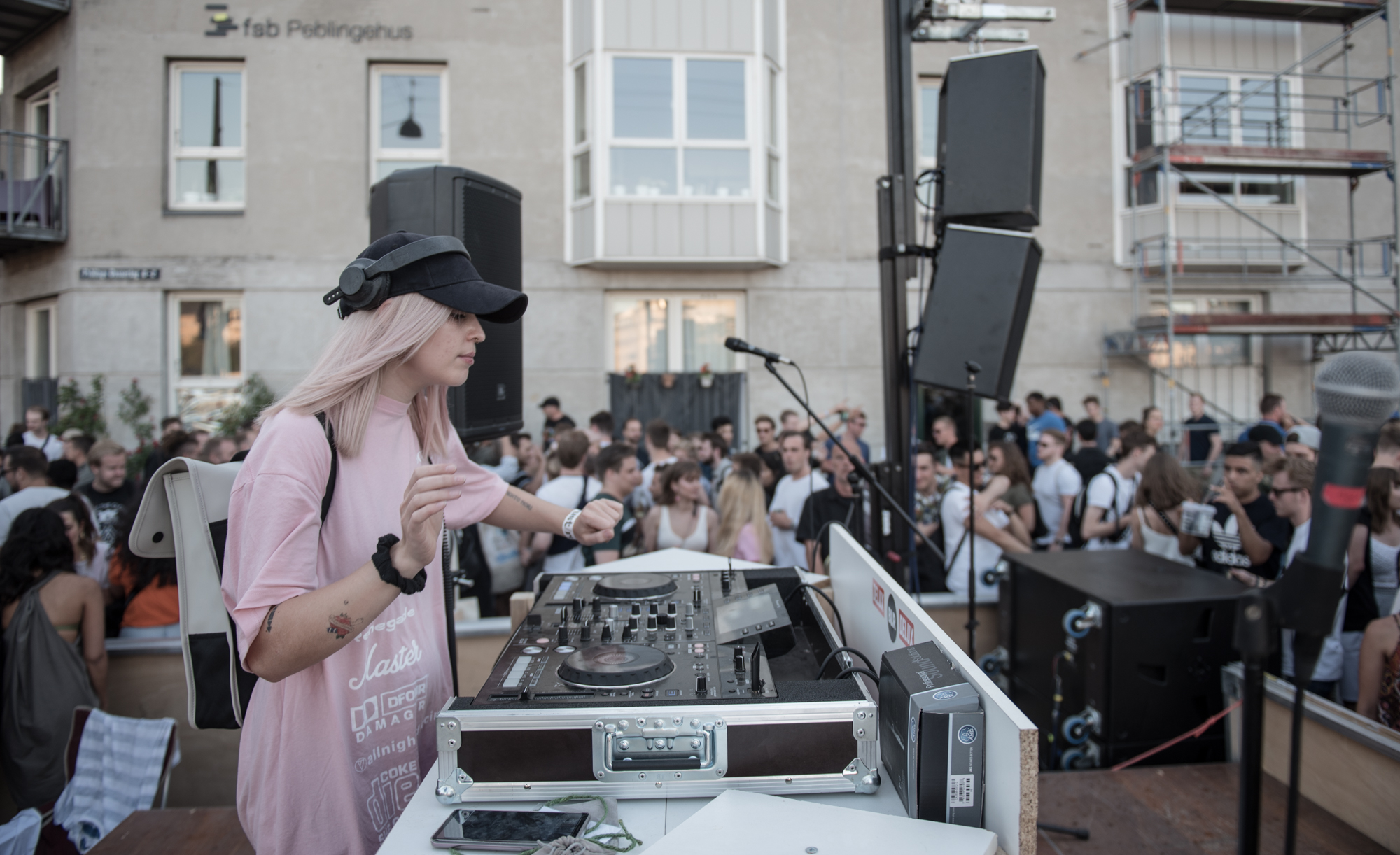 A DJ with pink hair and a baseball cap mixes music on a console at an outdoor event. She is surrounded by a crowd of people enjoying the performance. The background shows an urban setting with buildings and scaffolding.