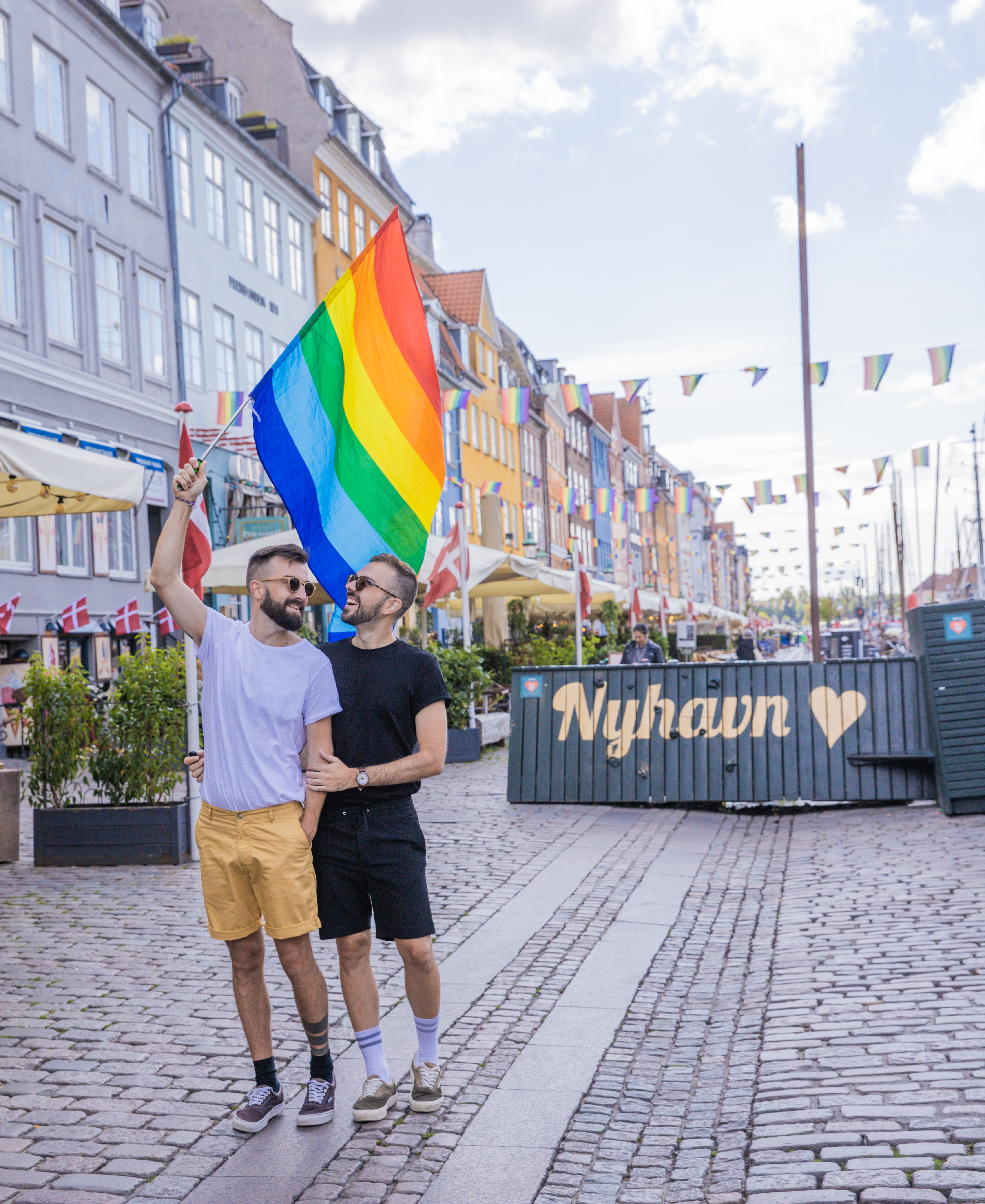 Two people walk happily in an open, cobblestone street, one holding a rainbow flag. Colorful buildings and triangular flags line the street. A container in the background reads Nyhavn with a heart symbol.