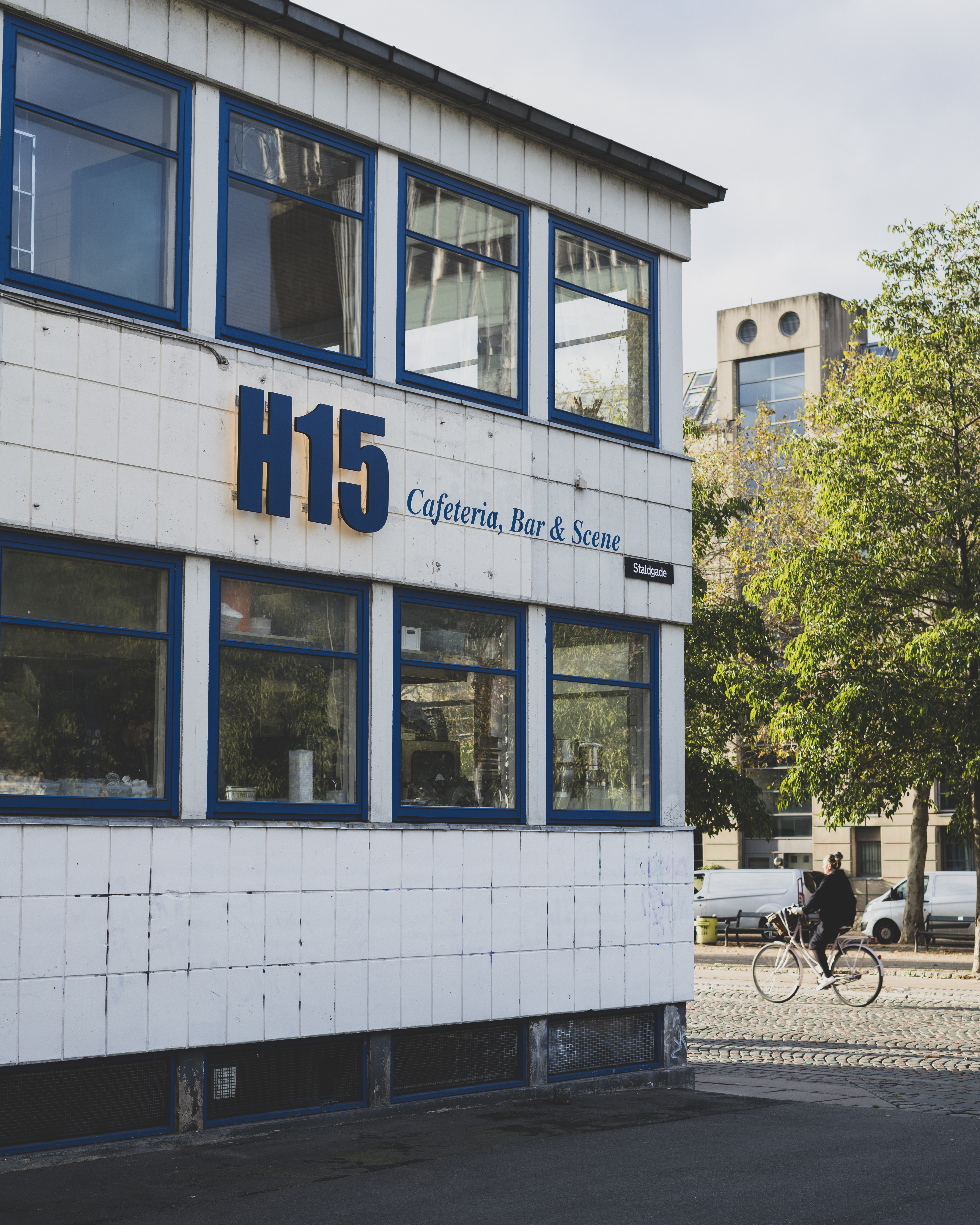 Street view of a building with a sign reading H15 Cafeteria, Bar & Scene. The facade is white with blue window frames. A person on a bicycle is passing by on the cobblestone street, and trees are visible in the background.