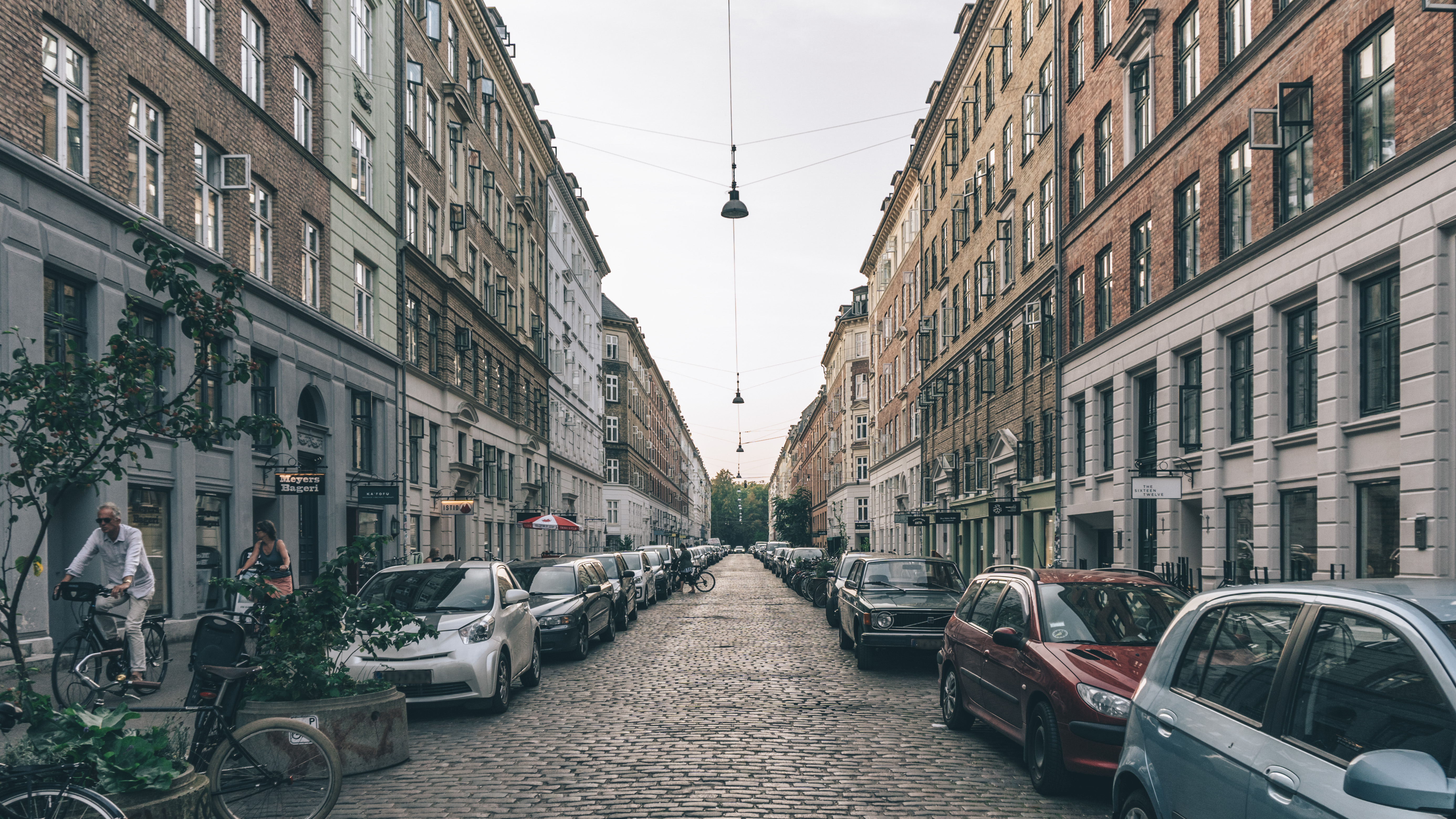 A cobblestone street lined with parked cars and tall, classical buildings on both sides. Lampposts hang over the street, and a few people are walking or cycling. The sky is overcast, creating a calm, urban atmosphere.