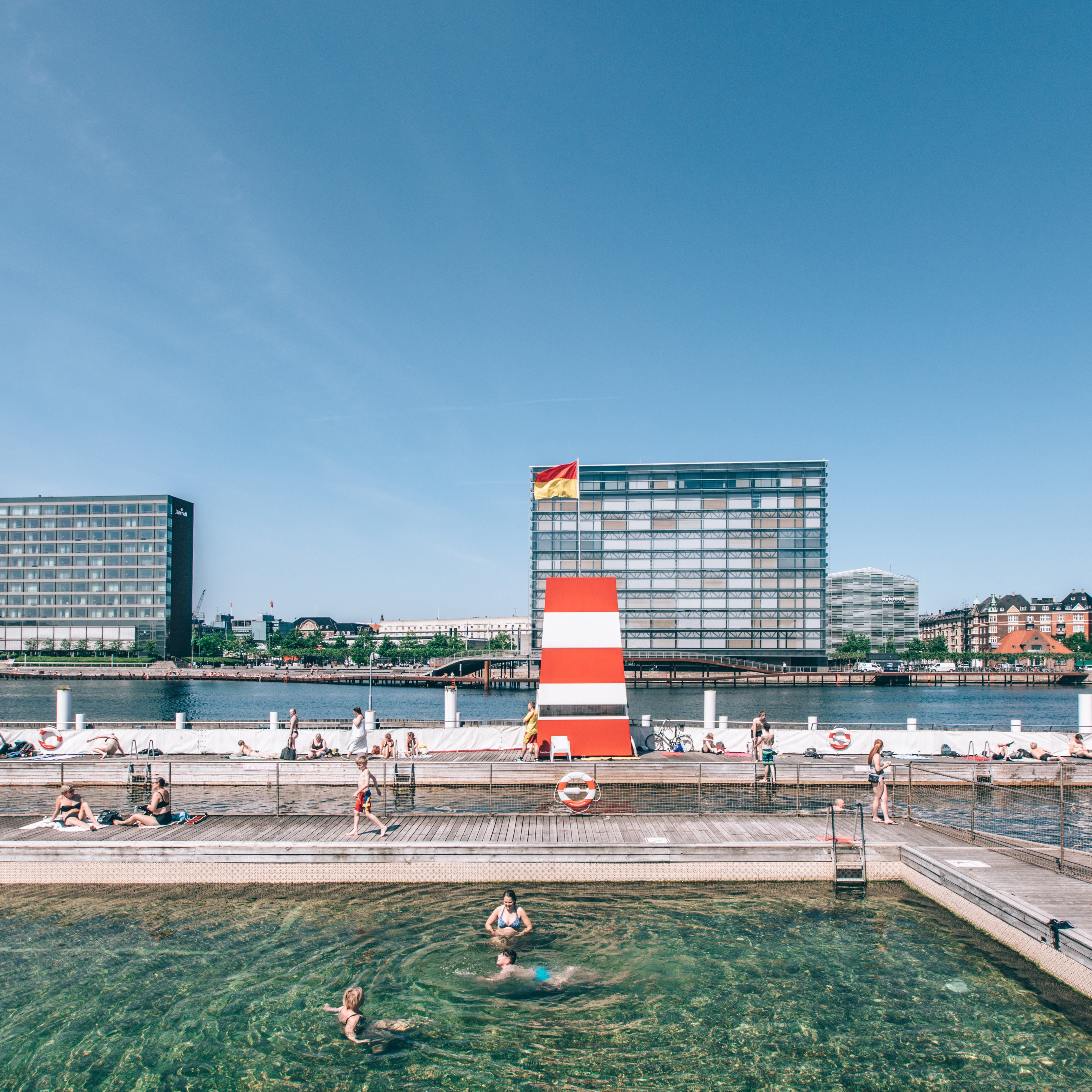 People swimming and sunbathing at a waterfront area with a wooden deck. A tall, striped structure stands at the waters edge. Modern buildings and a clear blue sky are in the background.