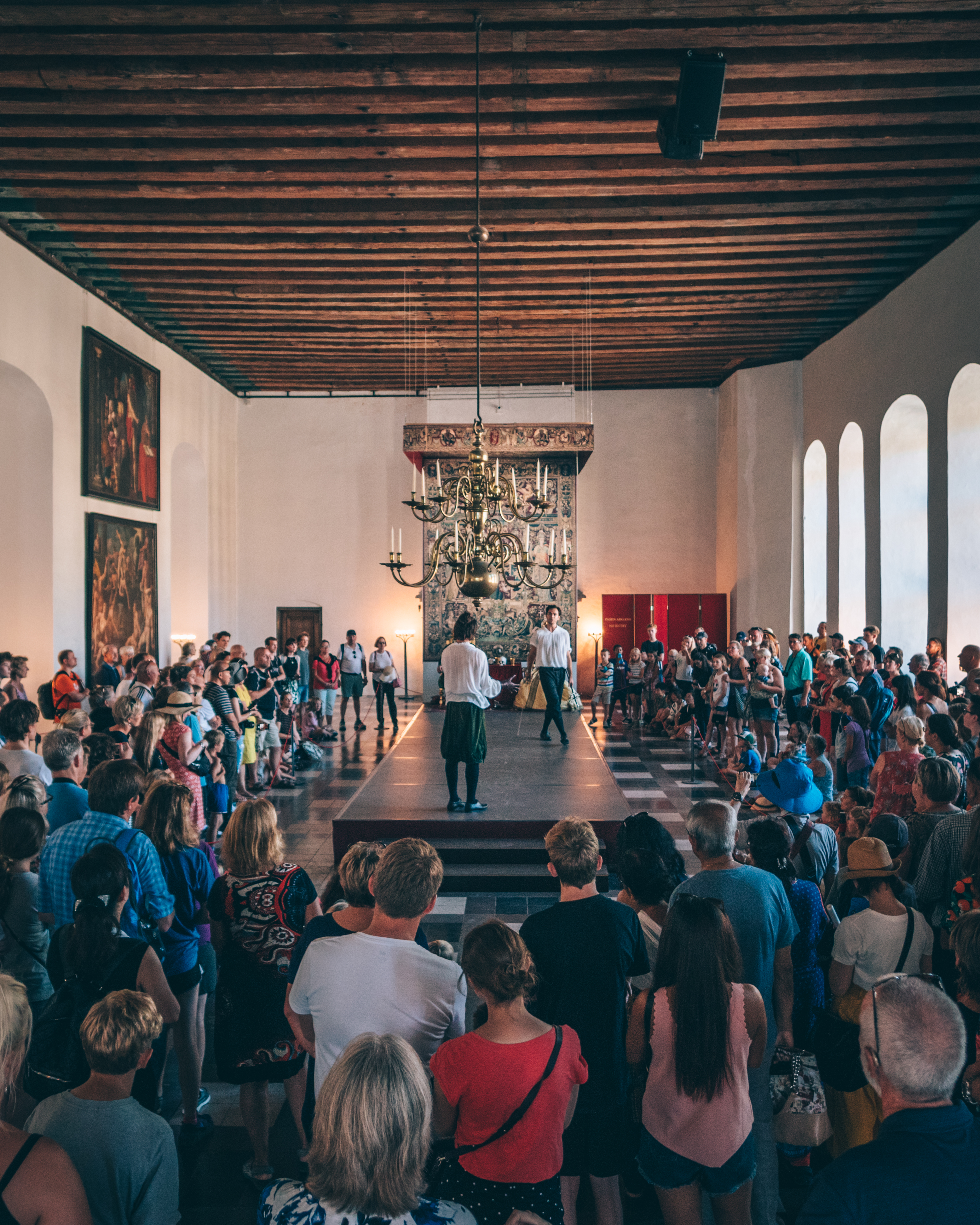 A large group of people gathers in a historic hall with high wooden ceilings and chandeliers, attentively watching two performers on a central platform. The walls are adorned with paintings, and the room has arched windows.