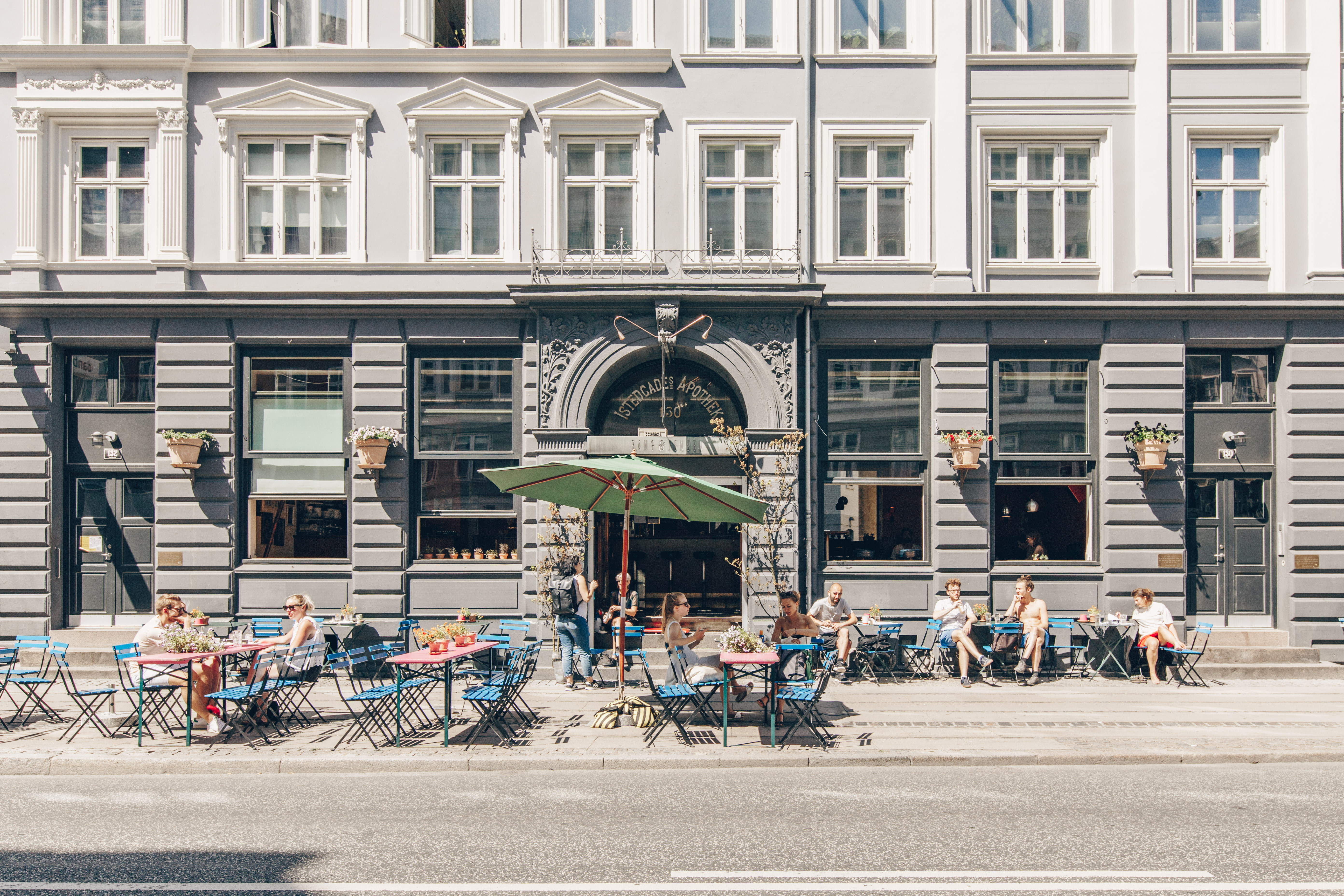 Street view of an outdoor café set against a historic building. People are seated at colorful tables under a large green umbrella, enjoying a sunny day. The building features ornate architecture, large windows, and small planters.