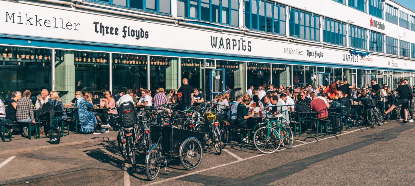 People enjoy outdoor dining at tables in front of a building featuring signs for Mikkeller, Three Floyds, and WarPigs. Many bicycles are parked on the sidewalk nearby. Its a bright, sunny day.