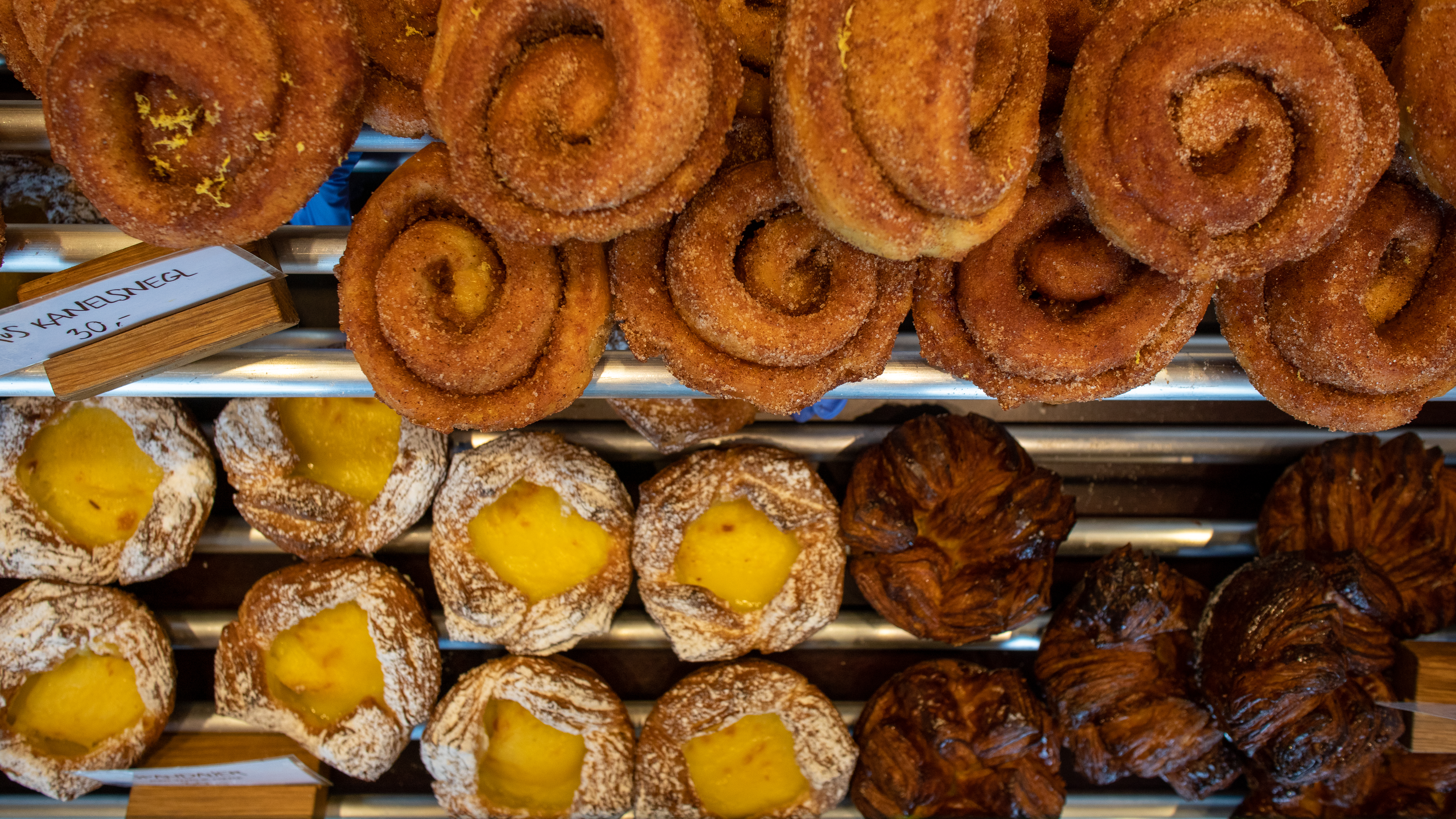 A variety of pastries on display: cinnamon sugar swirls on top, cream-filled pastries dusted with powdered sugar in the middle, and dark, flaky pastries at the bottom. Small price signs are visible beside the pastries.