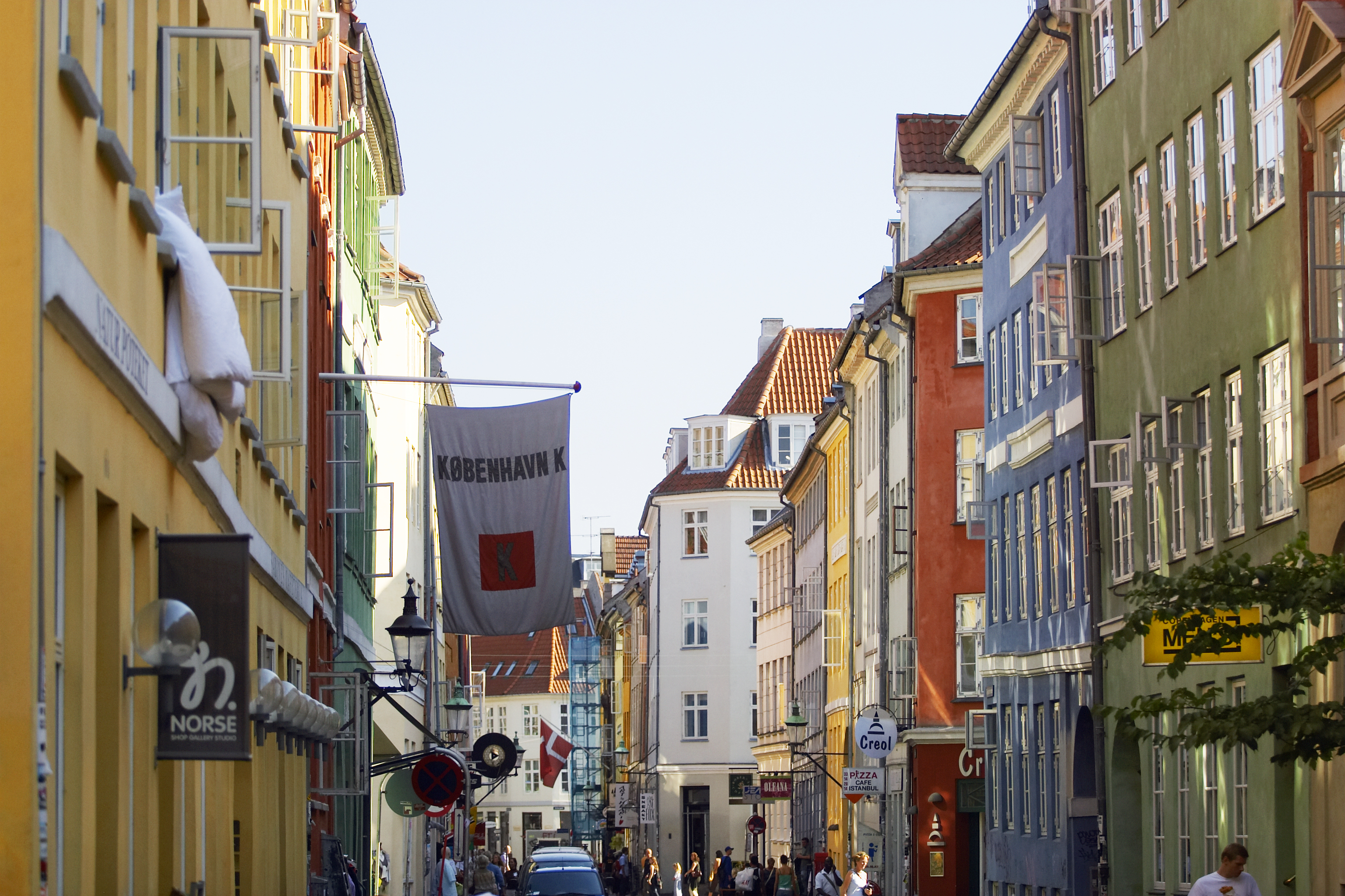 A street in Copenhagen with colorful buildings lining both sides. People are walking and cycling, and cars are parked along the street. A flag with København K hangs across the street. The sky is clear and blue.