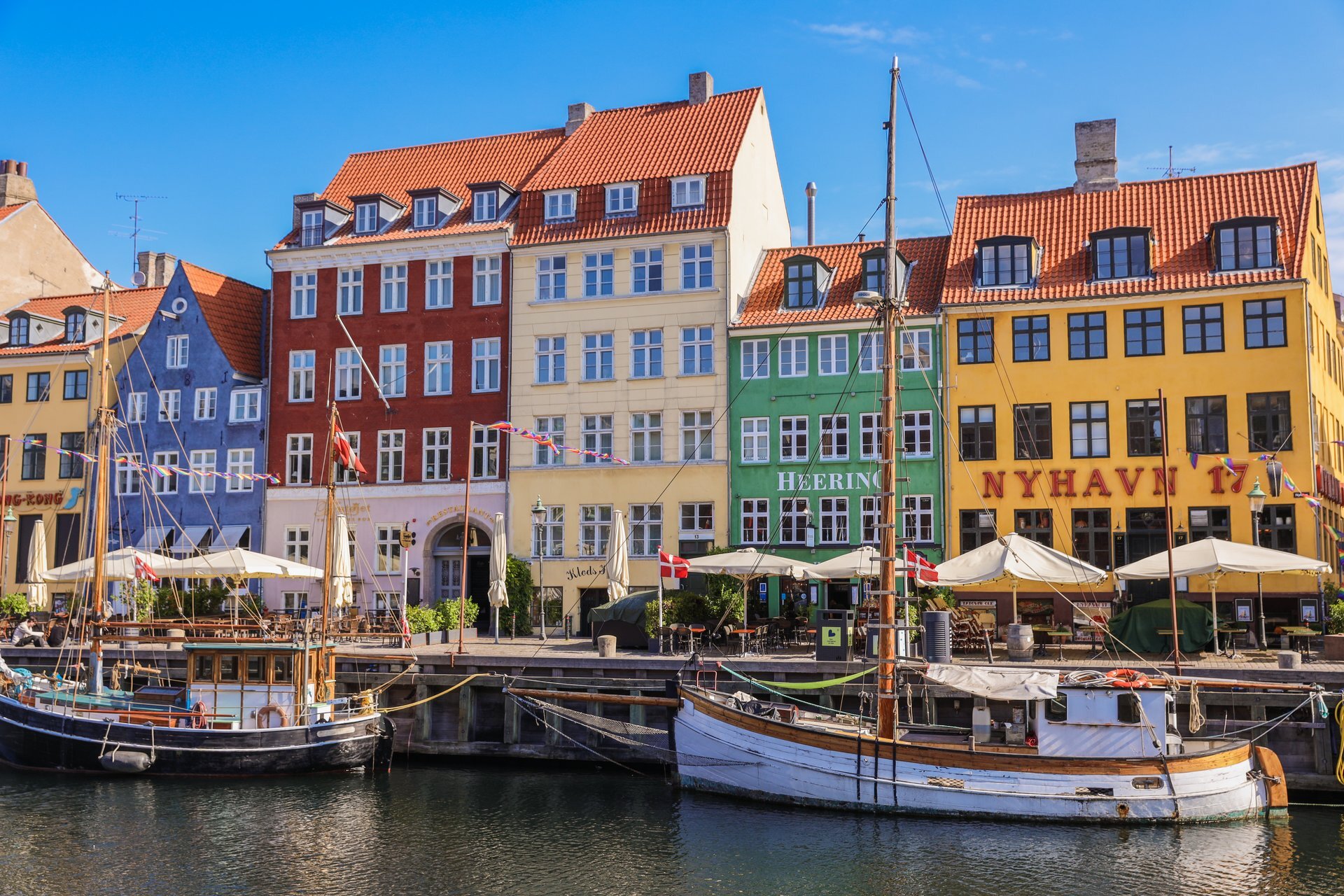 Colorful historic buildings line the Nyhavn waterfront in Copenhagen, Denmark. The structures are painted in red, yellow, and green, with outdoor cafes and sailing boats docked along the canal under a clear blue sky.