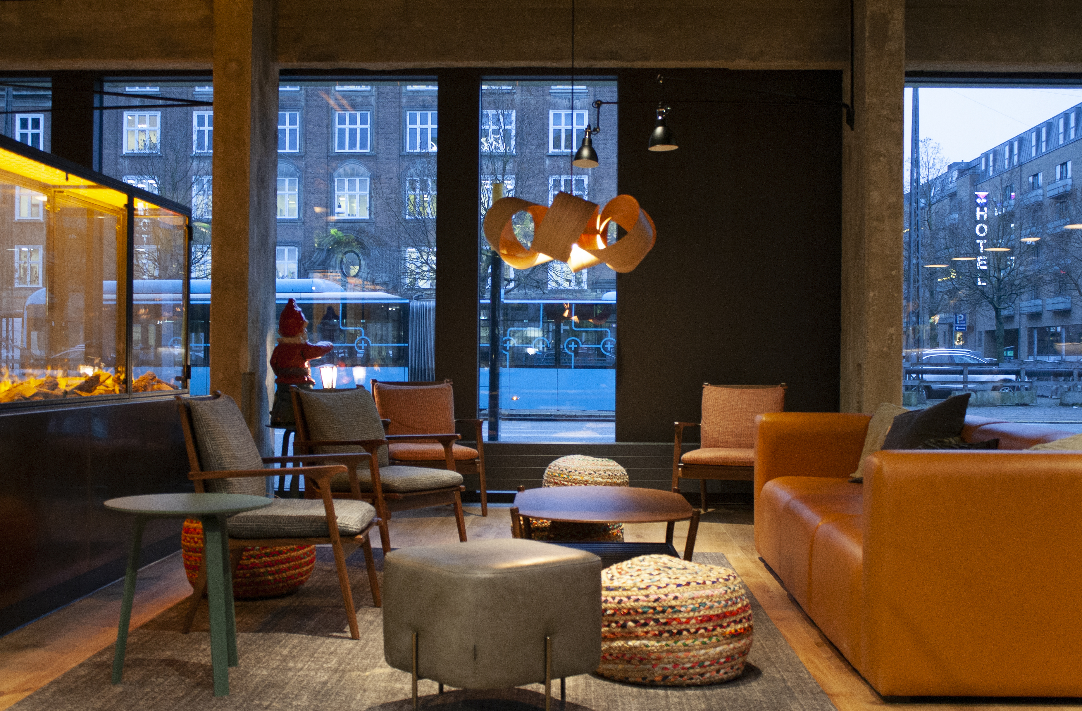 Modern lounge area with large windows, showcasing streetscape outside with a moving bus. Features leather and fabric chairs, a leather sofa, cozy lighting from a stylish pendant lamp, and a fireplace on the left. Urban atmosphere with daytime city view.
