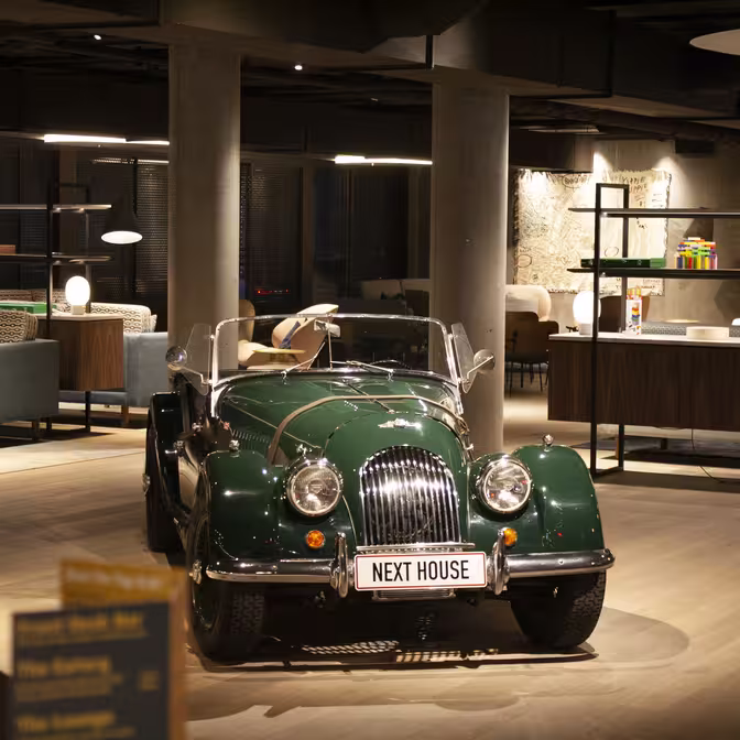 En grøn veteranbil med en "NEXT HOUSE"-nummerplade er udstillet indendørs i et moderne, stilfuldt rum. Indstillingen byder på moderne møbler, blød belysning og et slankt design, der skaber en blanding af klassisk og moderne æstetik.