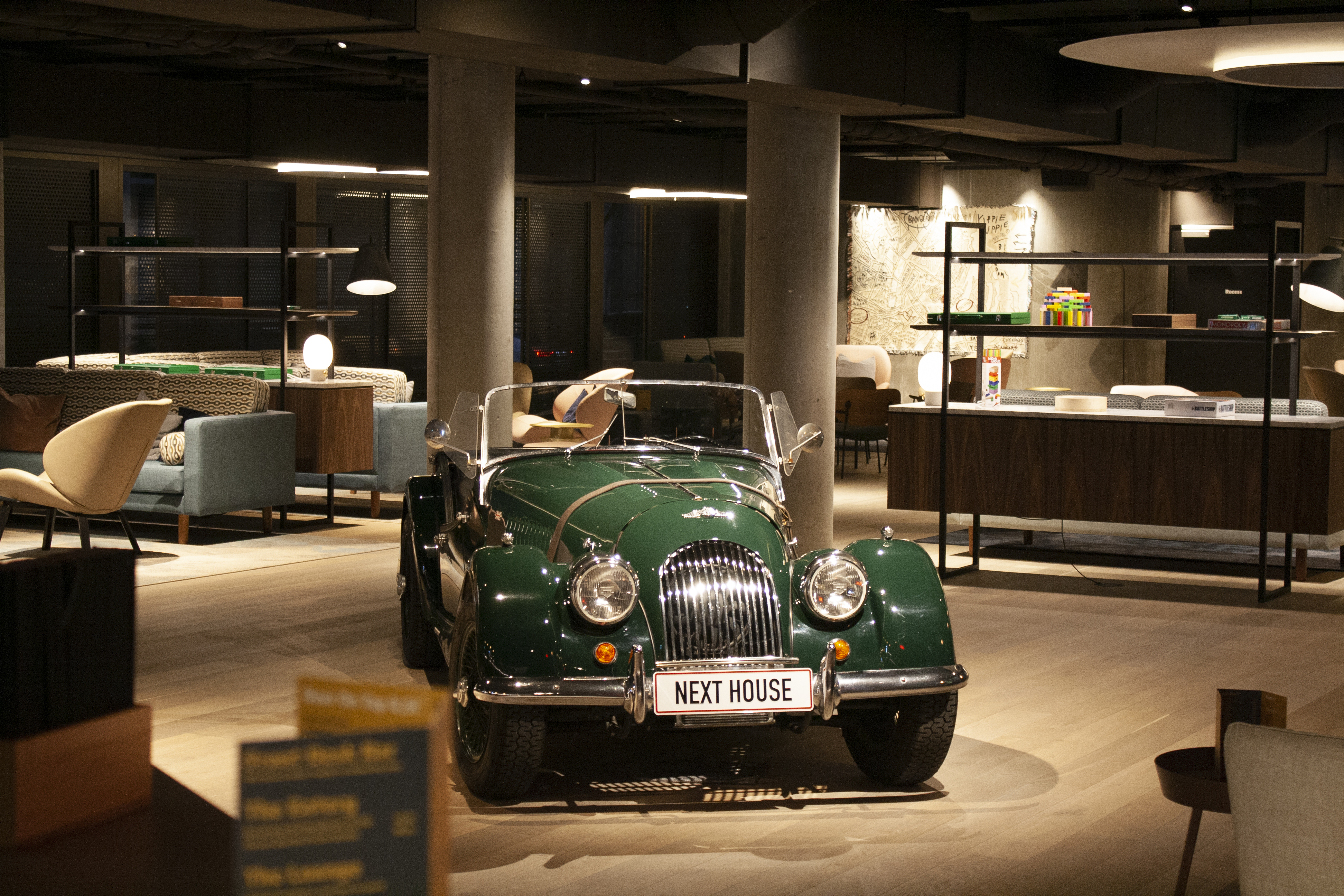 A vintage green convertible car is parked indoors on a wooden floor, surrounded by modern furniture including chairs and tables. The license plate reads NEXT HOUSE. The environment has an eclectic mix of classic and contemporary decor.