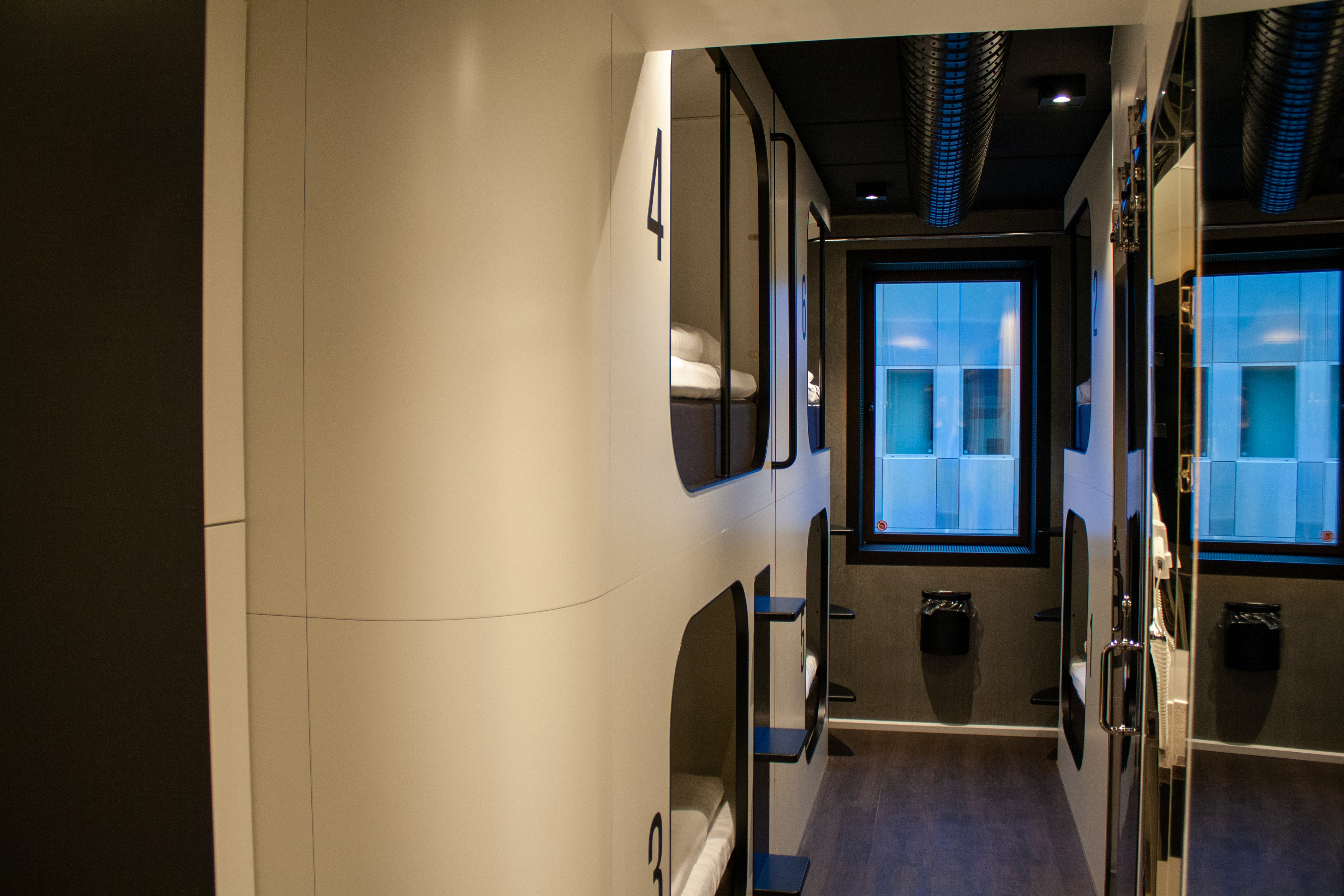 A modern capsule hotel interior with numbered pod beds stacked in a compact, sleek design. Each capsule has a small step and curtain for privacy, with a window at the end of the corridor providing natural light. Dark floor and ceiling panels complement the white pods.