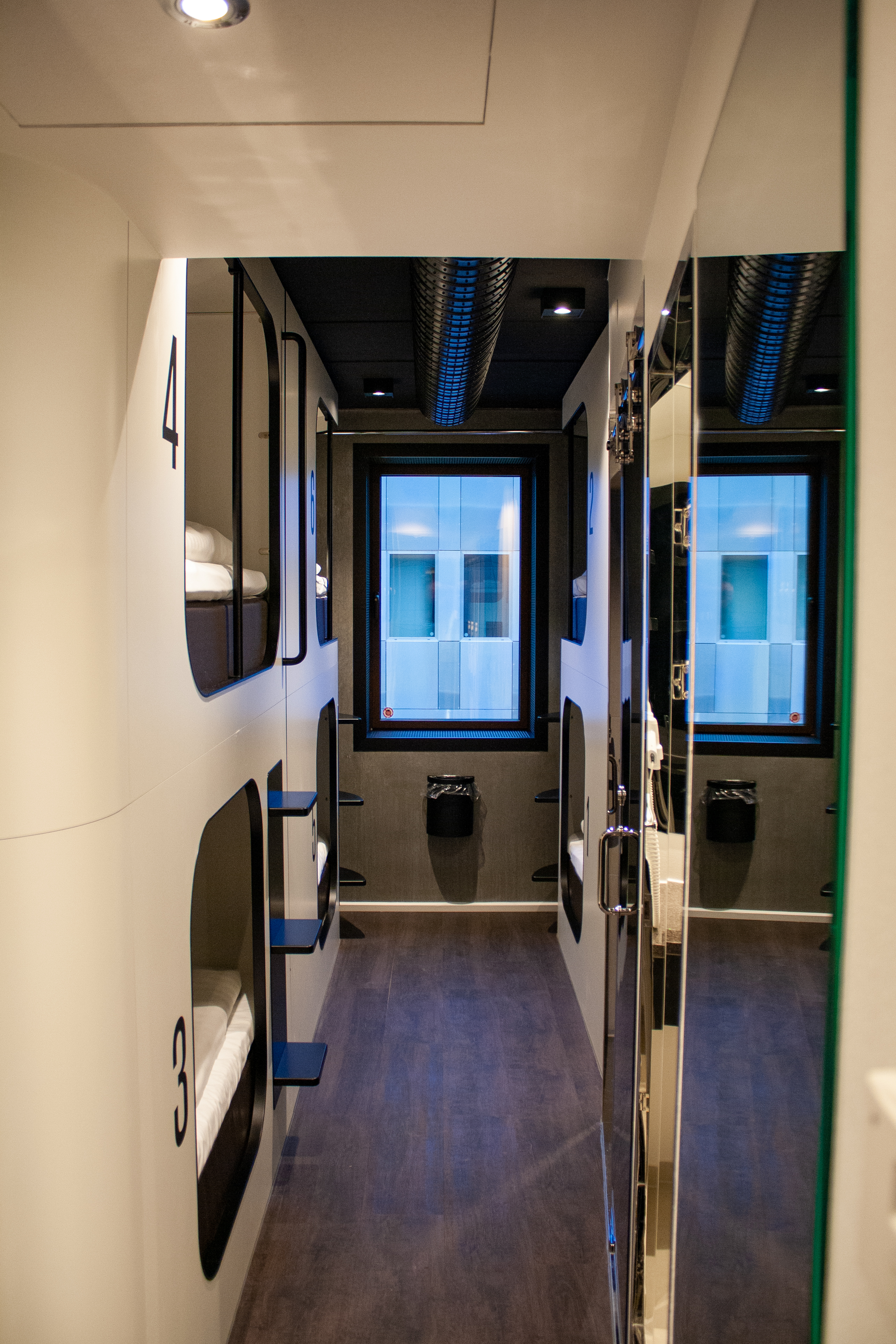 Interior of a modern capsule hotel room featuring two stacked sleeping pods with numbers 3 and 4. Each pod has a small ledge. The room is narrow with a window at the end, light wood flooring, and dark accents on the walls and ceiling.