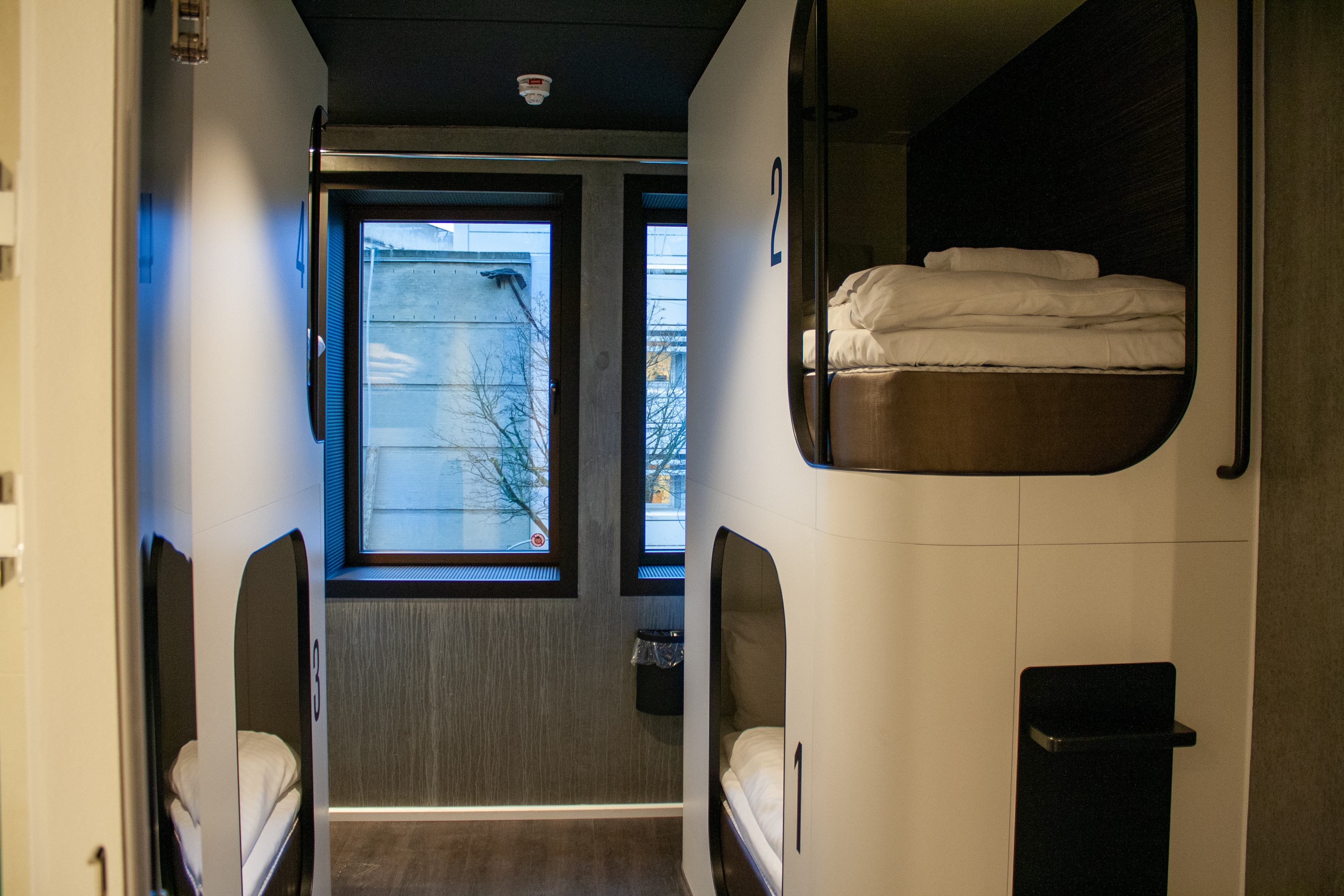 Interior of a modern capsule hotel room with two sets of bunk beds. Each capsule has neatly arranged bedding, individual lighting, and a number label. Large windows in the background allow natural light to enter.