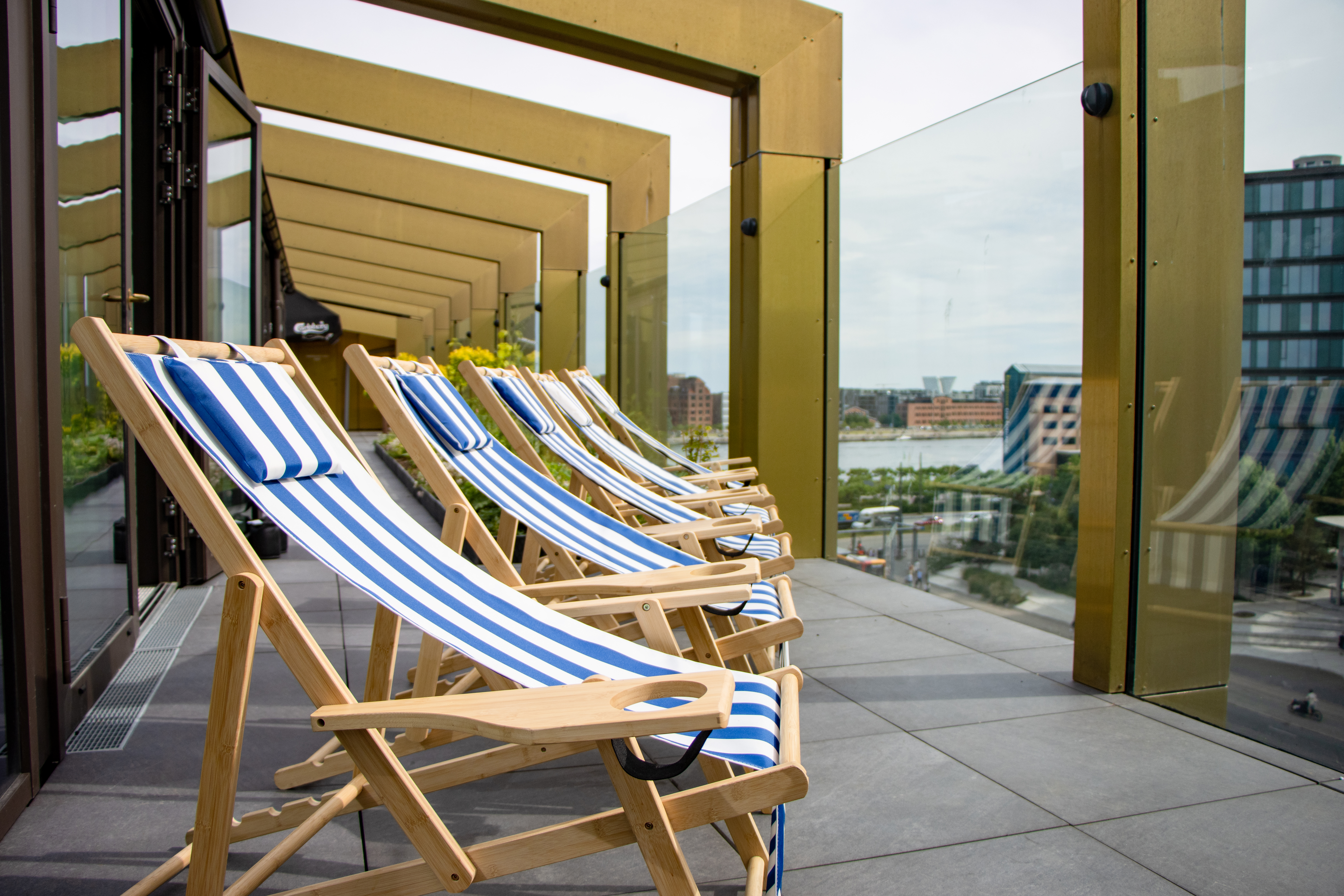 A row of striped deck chairs is arranged on a modern rooftop terrace with glass railings. The terrace overlooks an urban landscape, featuring buildings and a river in the distance under a cloudy sky.
