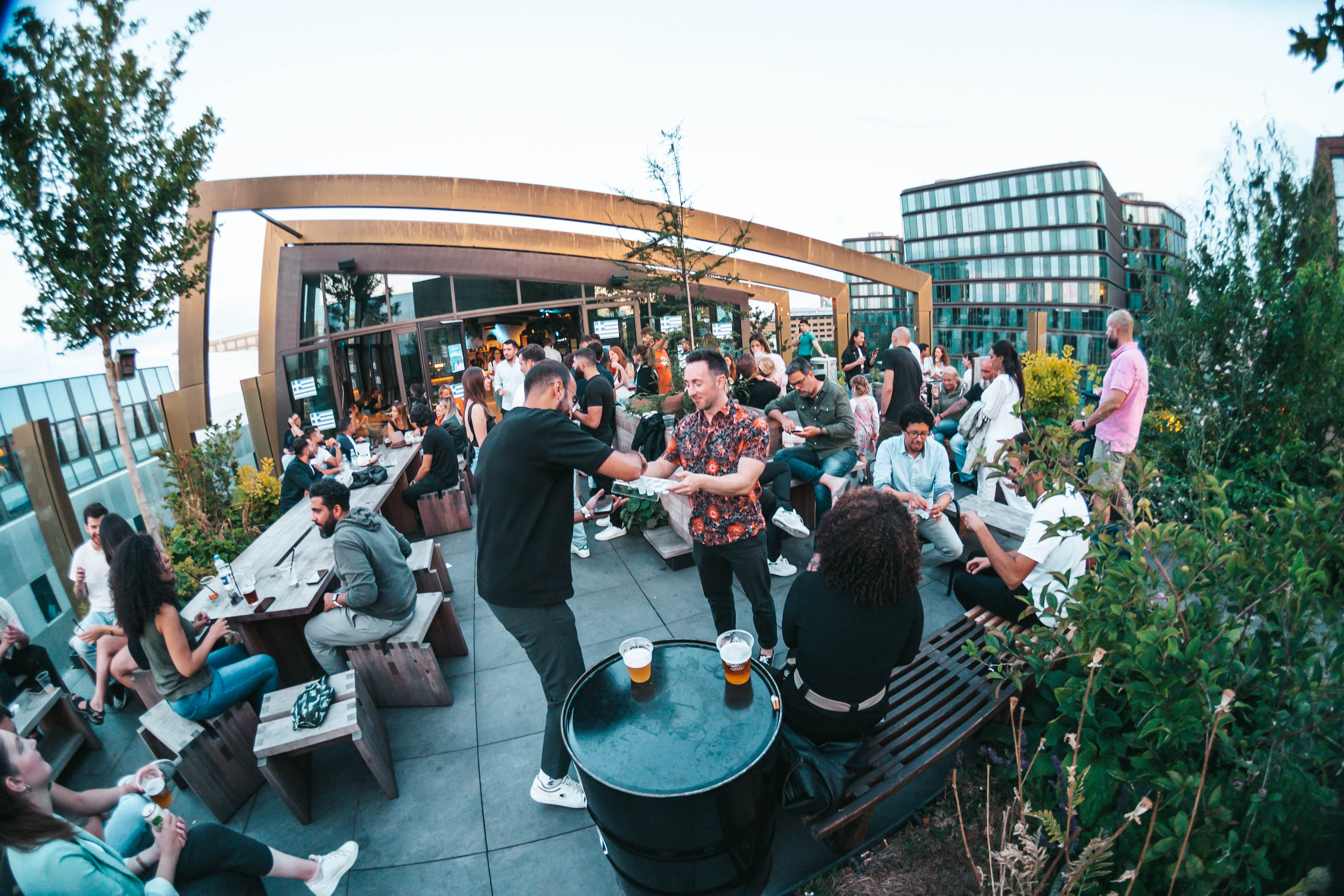 A lively rooftop bar scene with people socializing and enjoying drinks. Tables are scattered around with patrons seated and standing. Greenery decorates the area, and modern buildings are visible in the background.