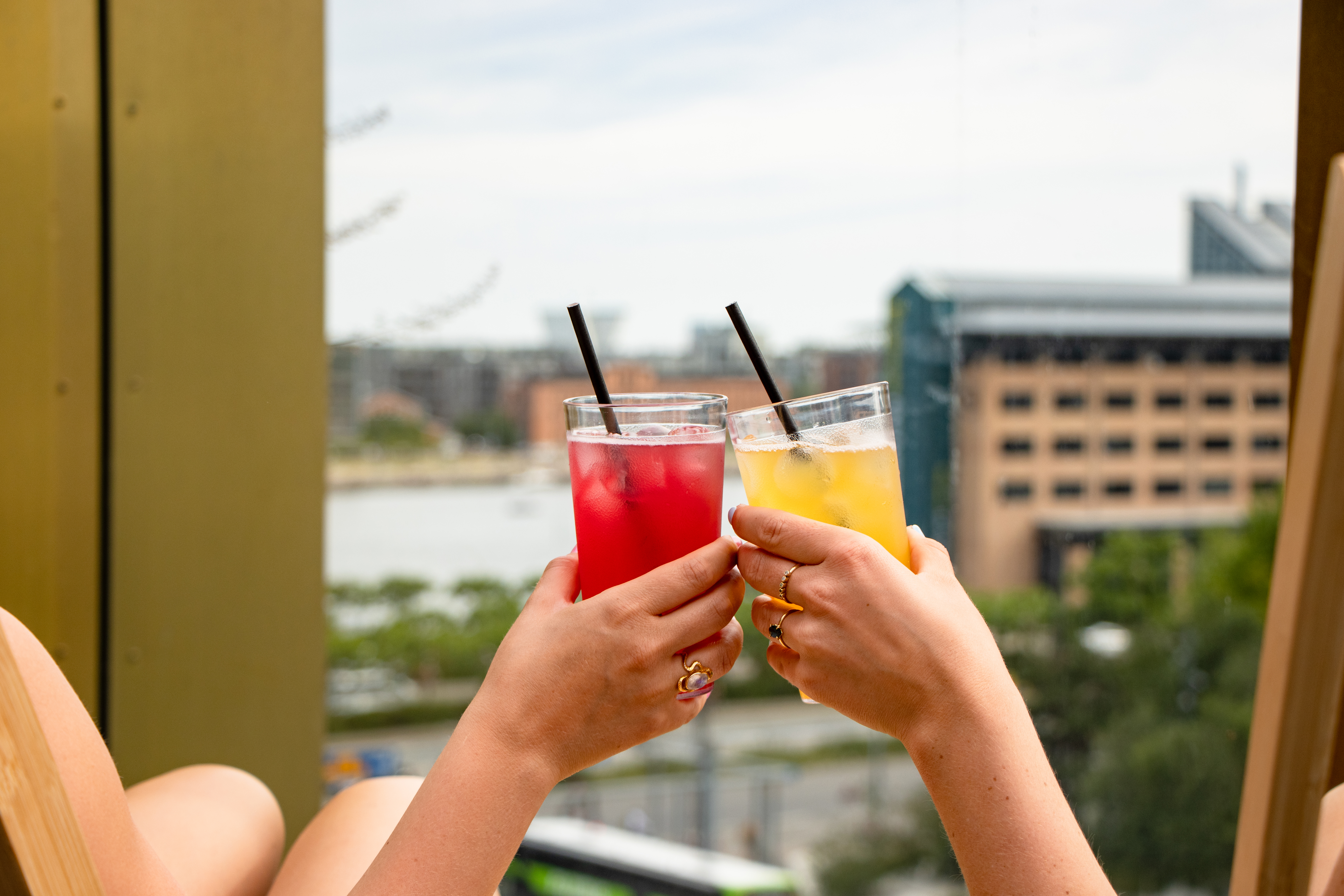 Two people clink glasses filled with colorful drinks, one red and one yellow, against a blurred backdrop of a cityscape and water. Both glasses have straws and ice cubes. The mood is relaxed and summery.