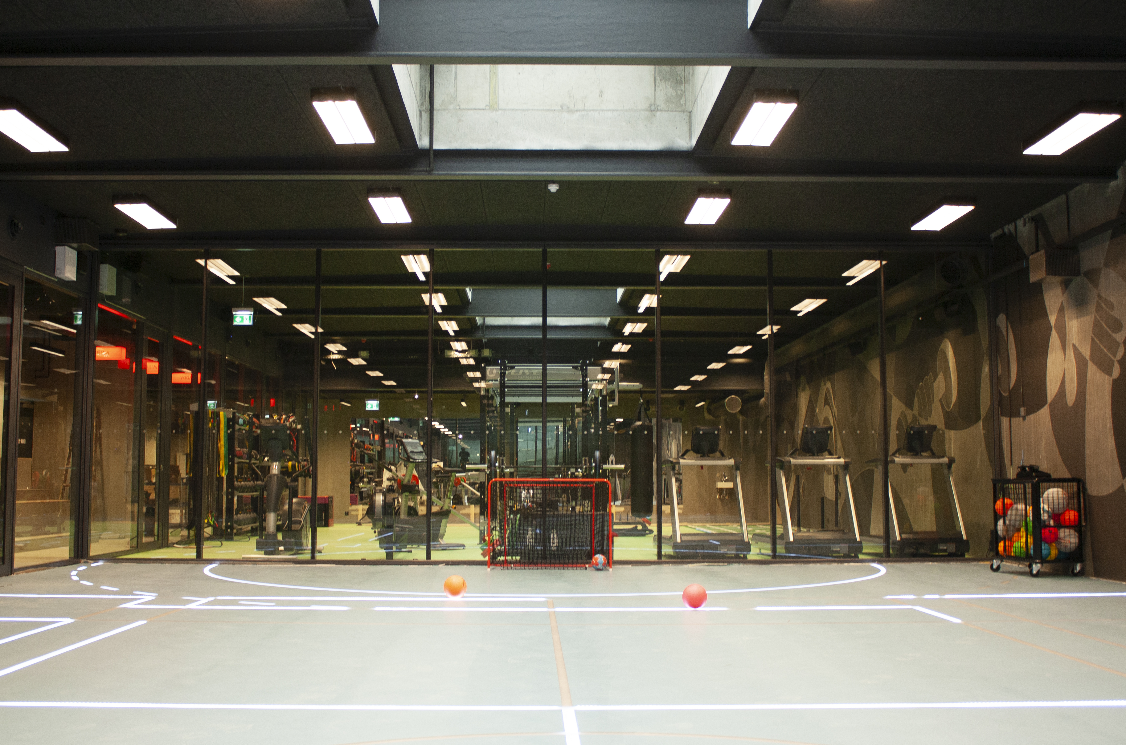 A modern gym with a glass wall revealing treadmills, exercise equipment, and a neon-lit interior. The foreground shows a sports field with two balls and a set of small hockey goals on the floor.