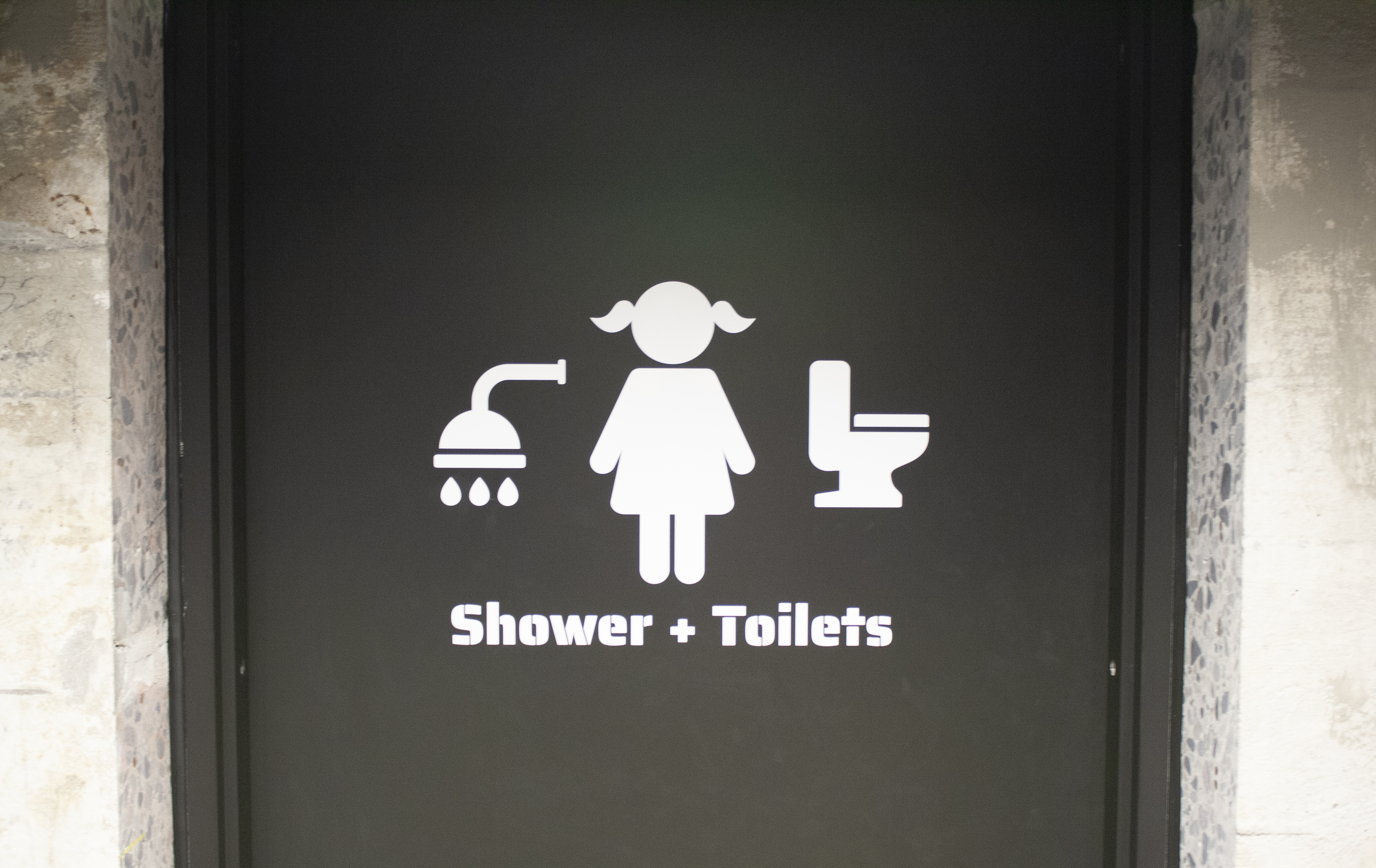 A black restroom door with white icons depicting a showerhead and toilet surrounding a person with short hair. Below the icons, text reads Shower + Toilets.