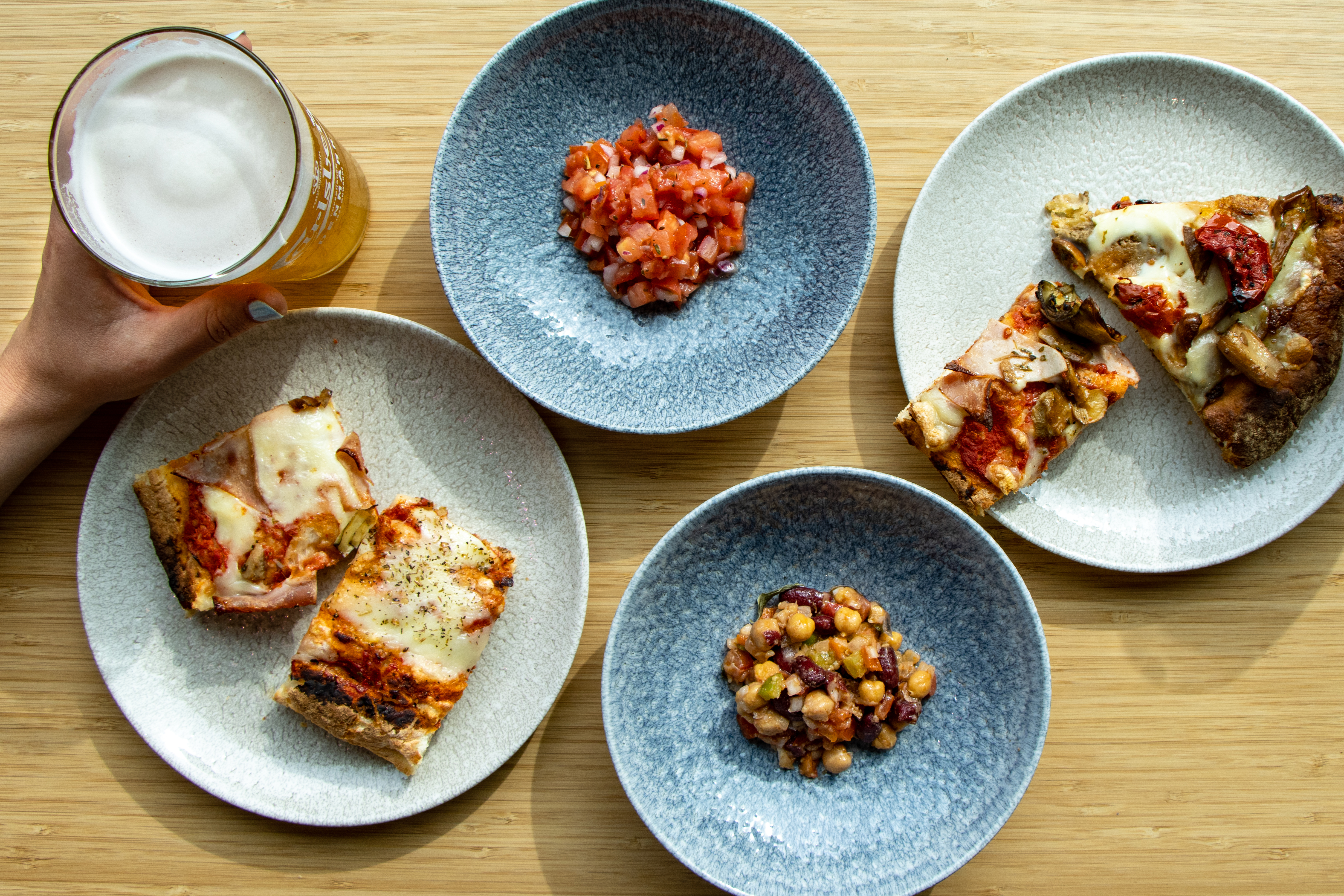 A hand holds a glass of beer next to four plates on a wooden surface. The plates contain different slices of pizza, diced tomatoes, and a chickpea salad, all served on blue-gray plates.