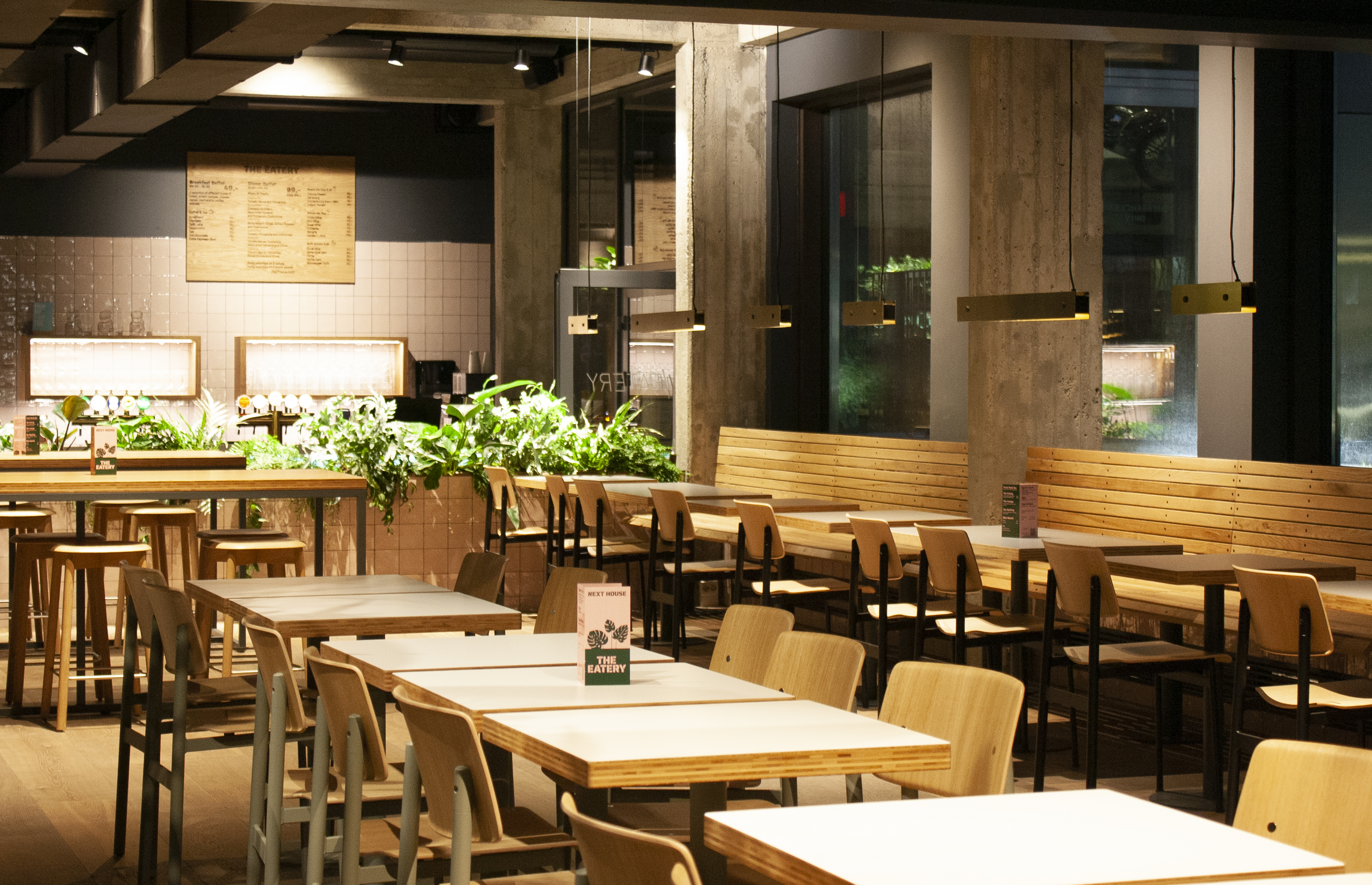 A modern café interior with wooden tables and chairs. The space features industrial design elements, large windows, and greenery along the walls. Soft, warm lighting creates a cozy atmosphere. Menu boards are visible in the background.