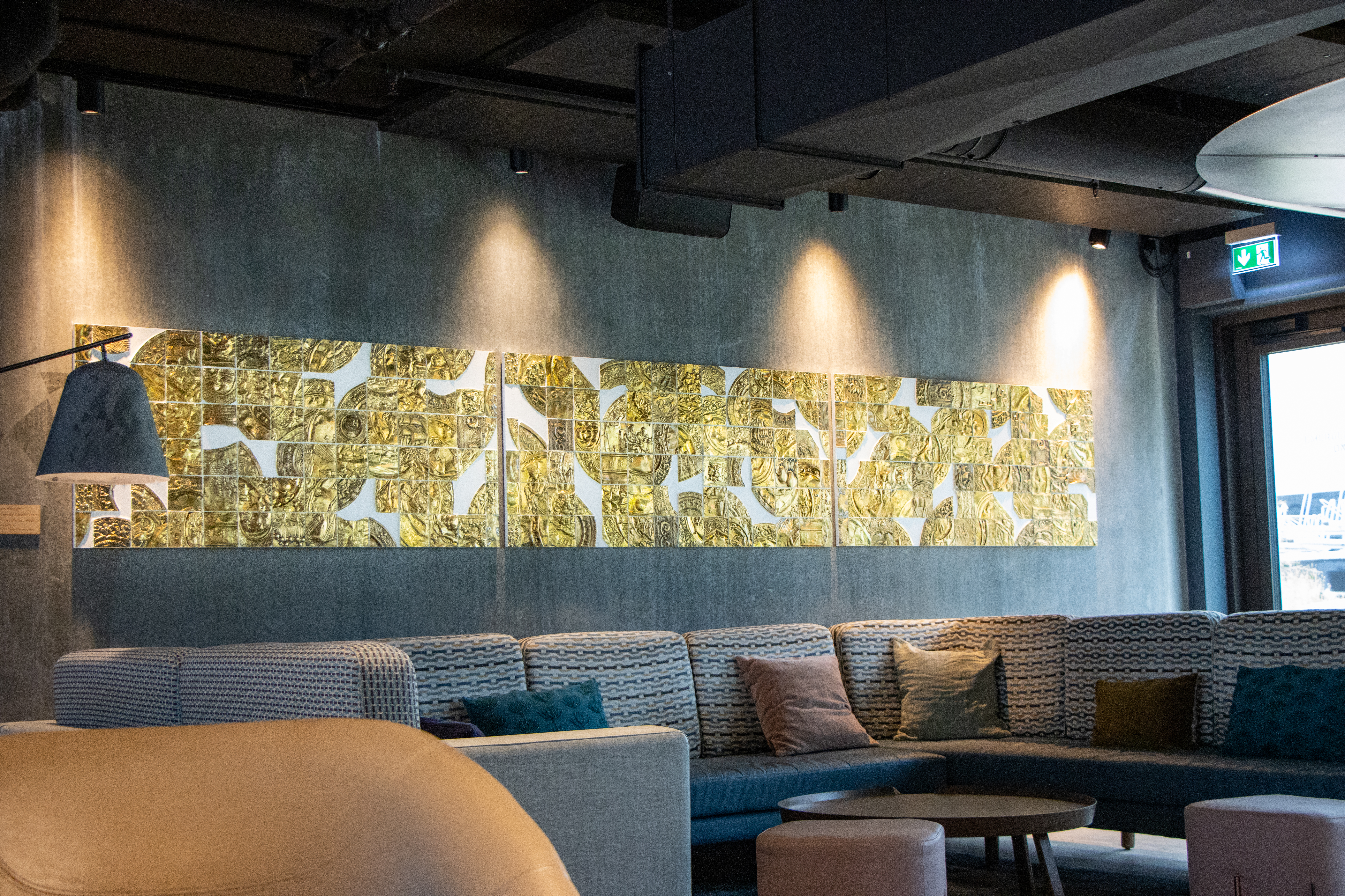 A modern lounge area with a long couch featuring patterned cushions. Above the couch, a colorful abstract art piece is illuminated on the wall. The room has dim lighting, a hanging lamp, and an emergency exit sign near a window.